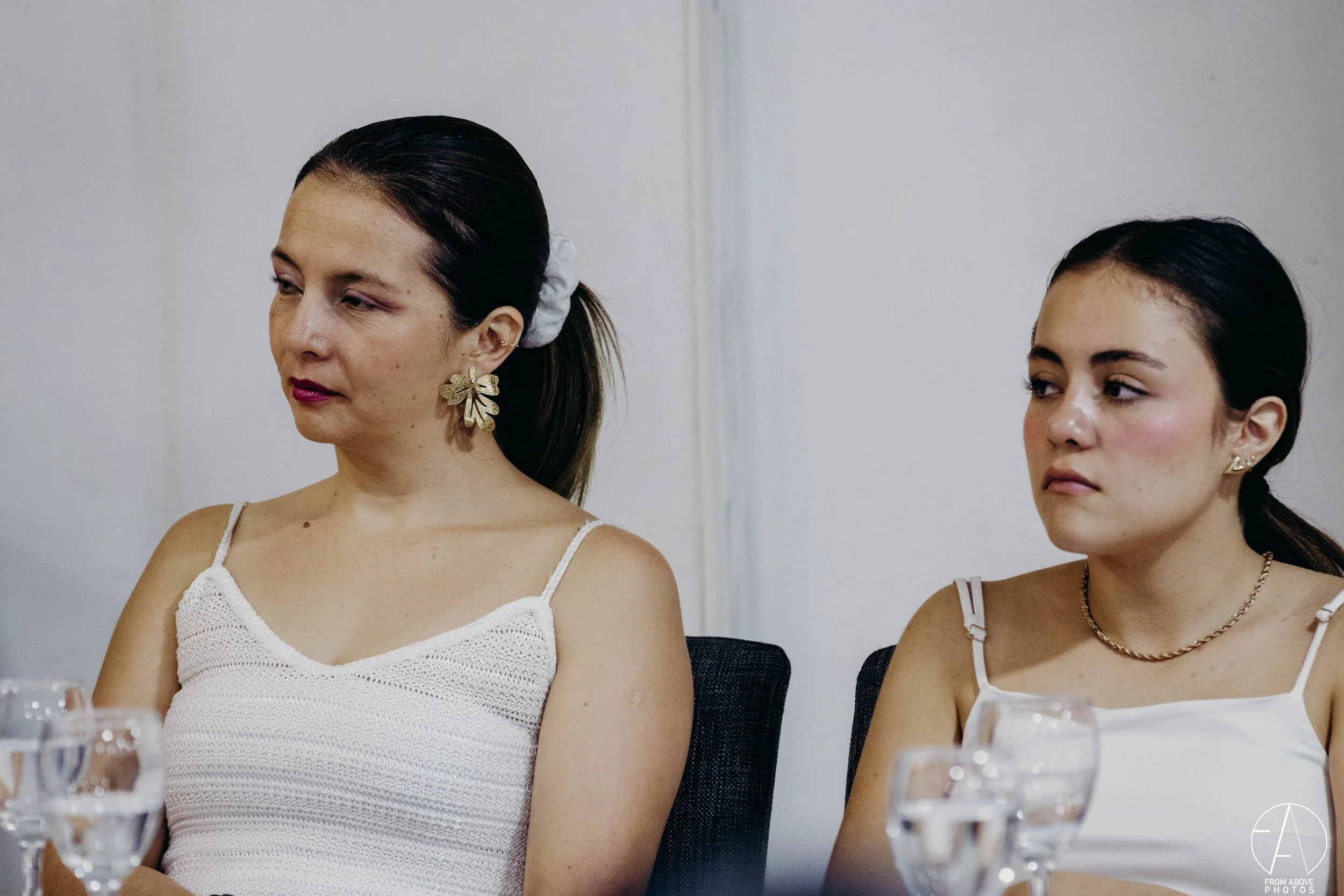 Dos mujeres sentadas en una mesa, una de ellas con expresión pensativa y la otra con expresión neutra, en un entorno interior.