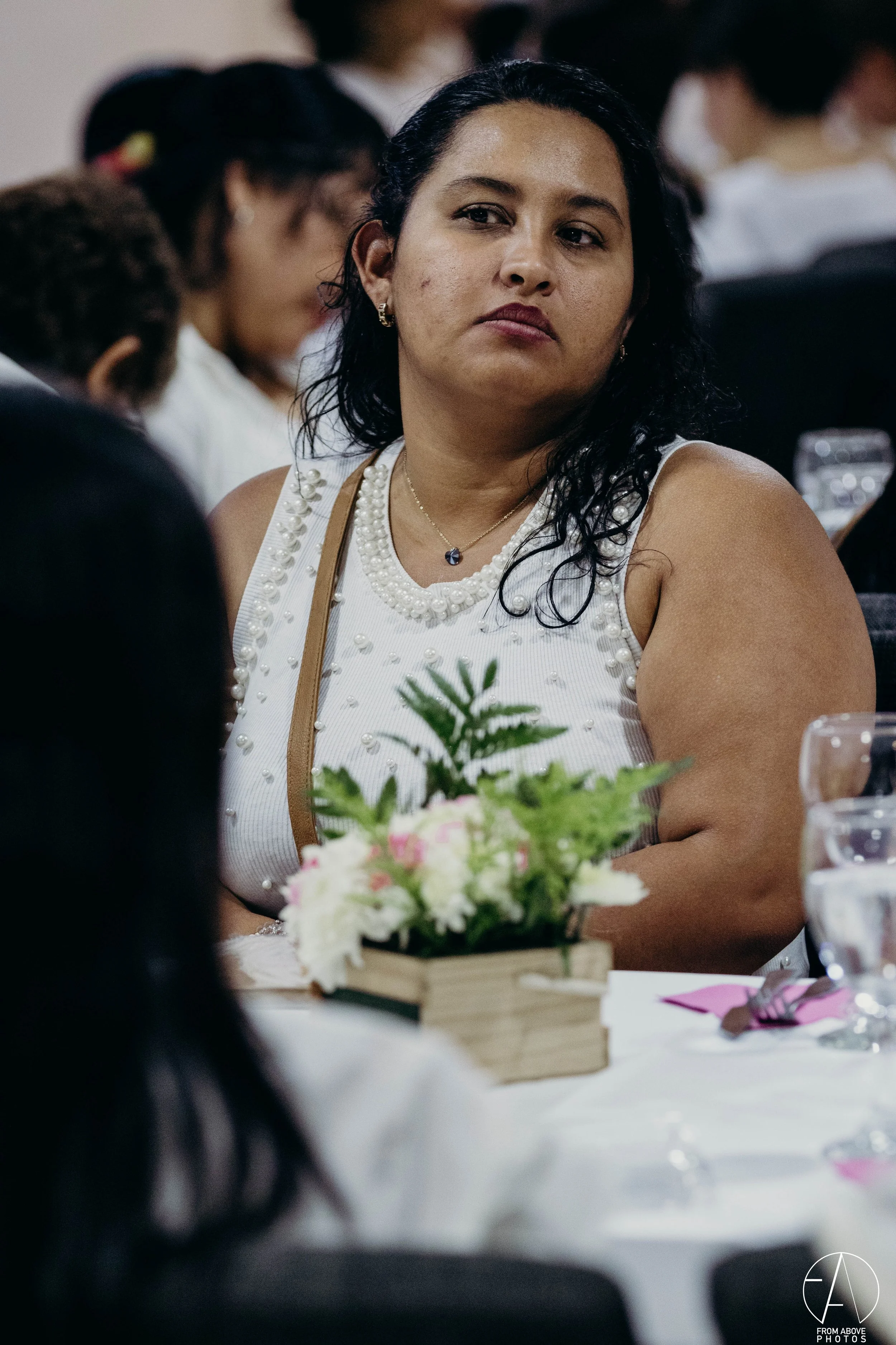 Mujer con maquillaje natural, vestida con blusa blanca con detalles de perlas, en una reunión social, rodeada de otras personas y decoraciones florales en la mesa.