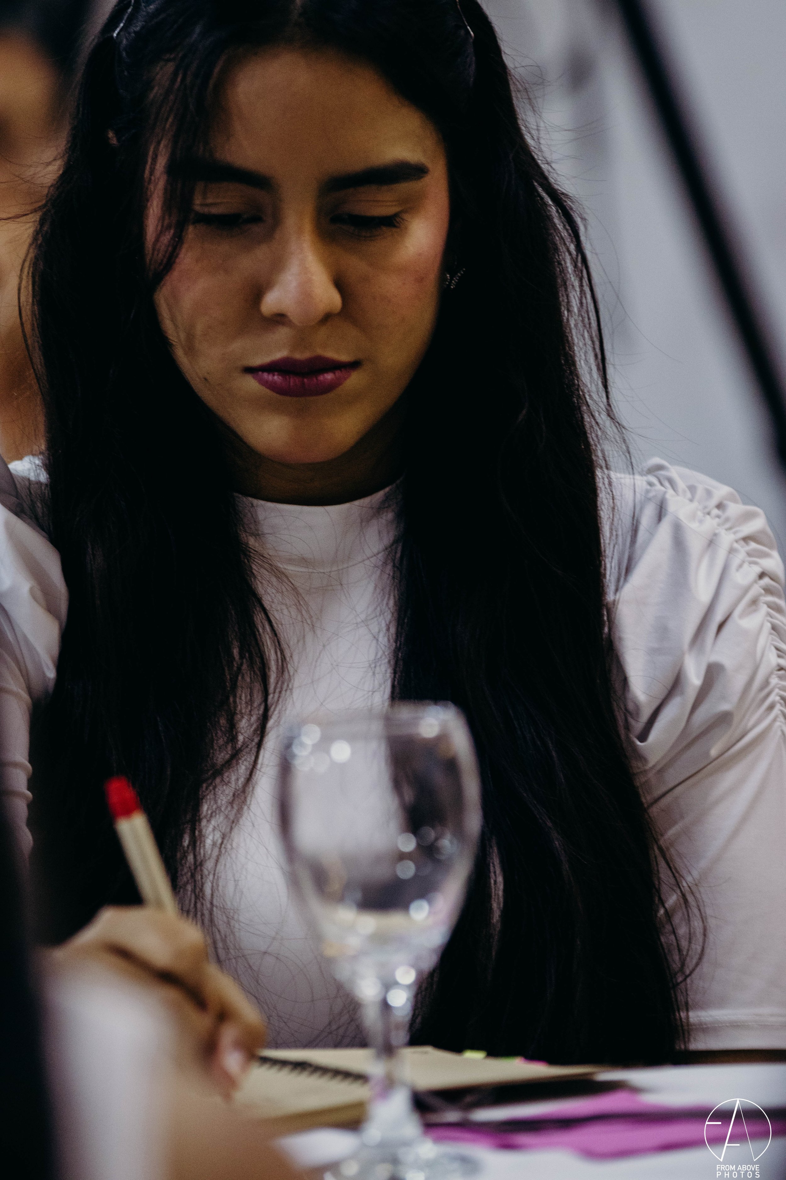 Una mujer de cabello largo y oscuro, con maquillaje de labios morados, escribe en un cuaderno con un bolígrafo rojo, en una mesa con una copa de vino tinto frente a ella.