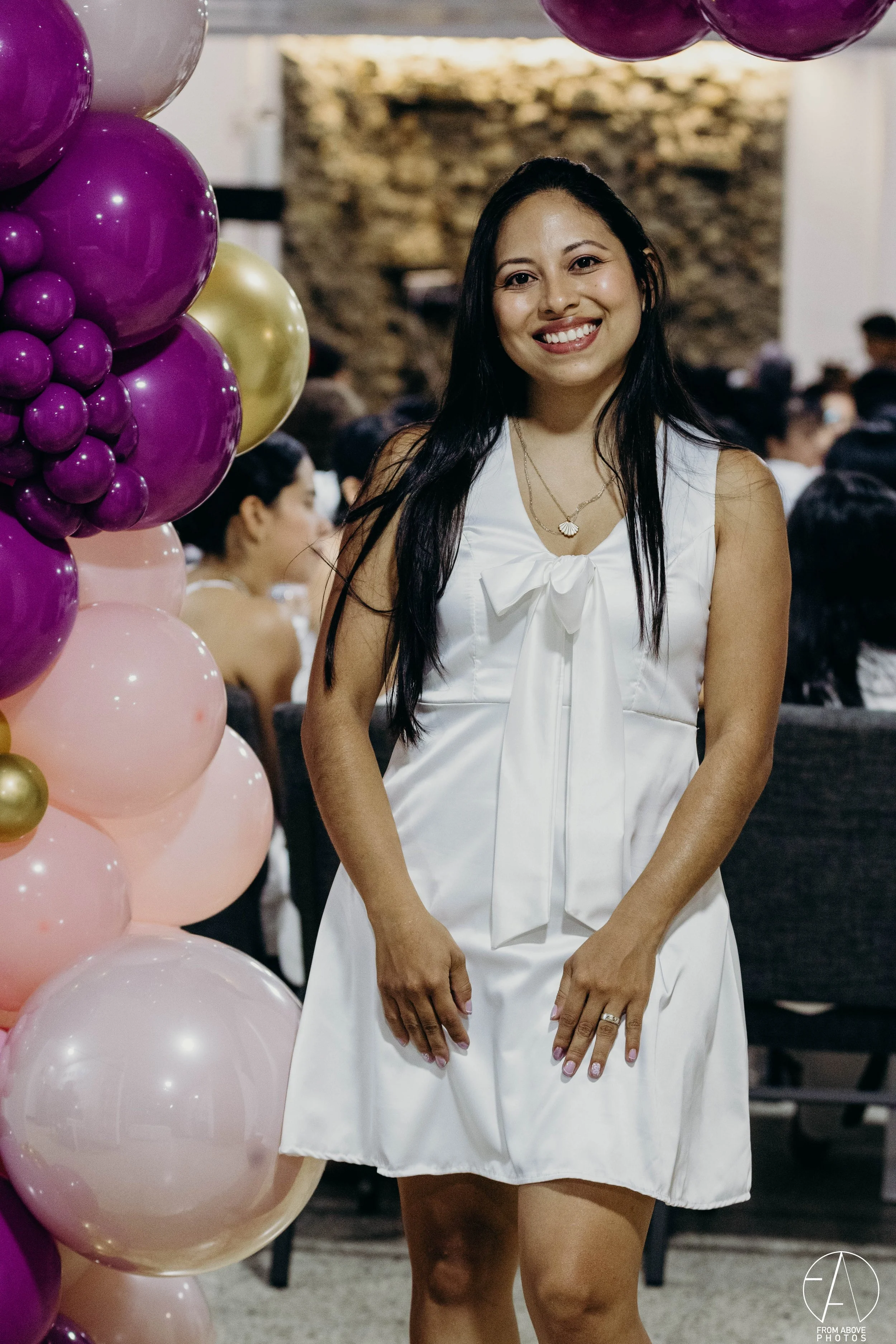 Mujer sonriendo en un evento con globos morados, rosados y dorados, vestida con un vestido blanco con lazo en el cuello.