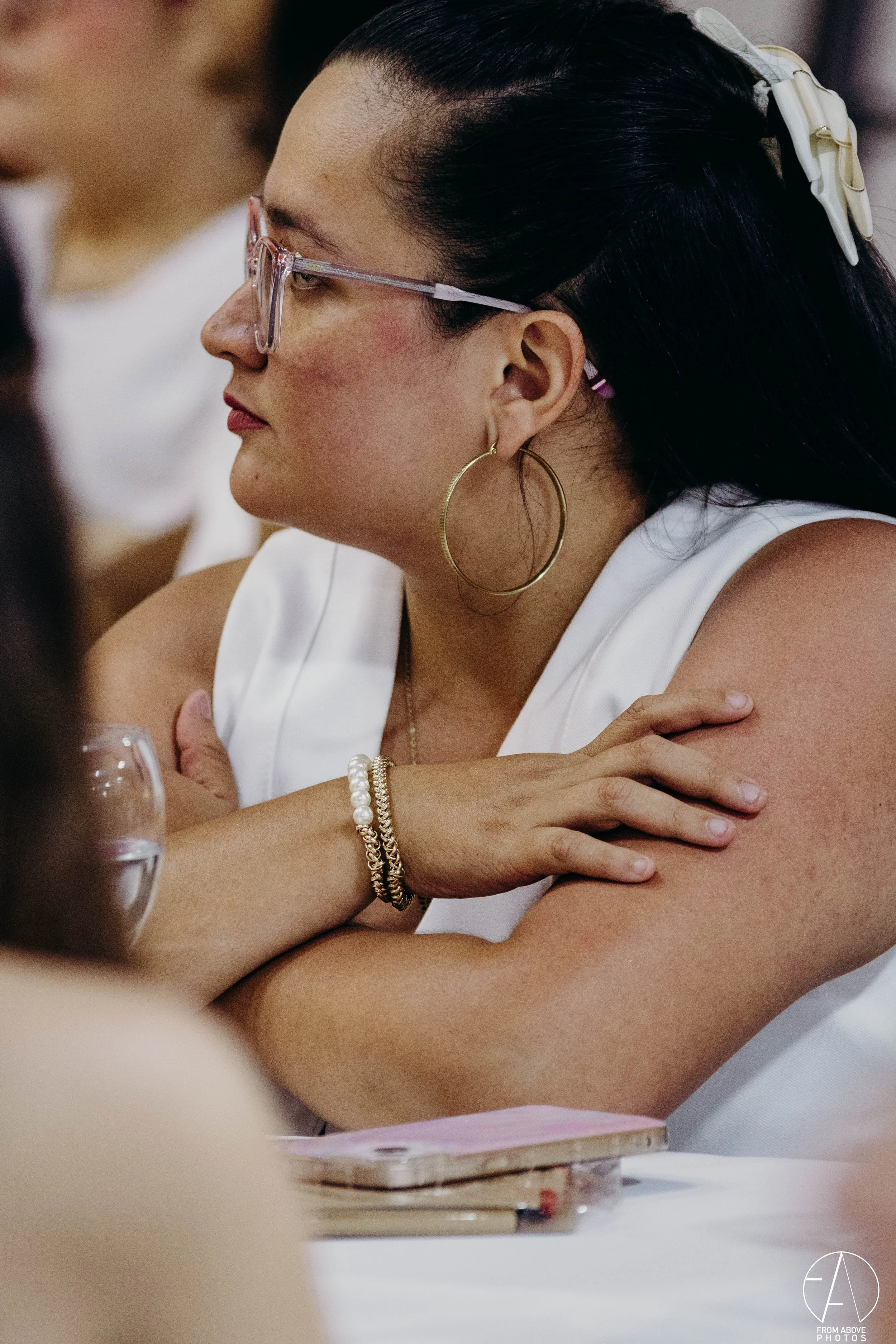 Una mujer con gafas transparentes, aretes grandes de aro dorados, peinado con una coleta y accesorios blancos, está sentada con los brazos cruzados en una mesa, con un fondo difuso