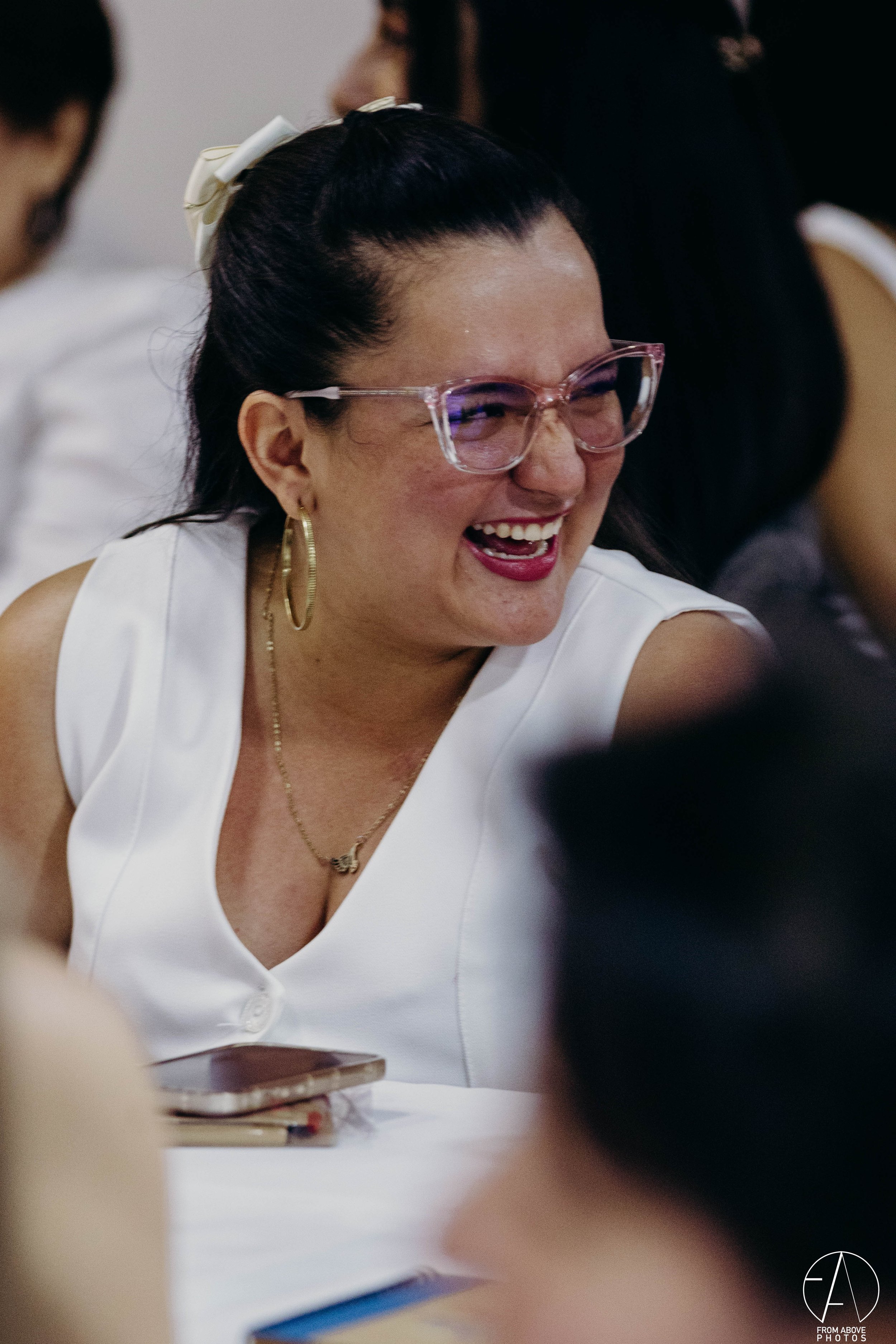 Mujer sonriendo con gafas grandes, peinado en cola de caballo con lazos, usando blusa blanca, en un ambiente social.