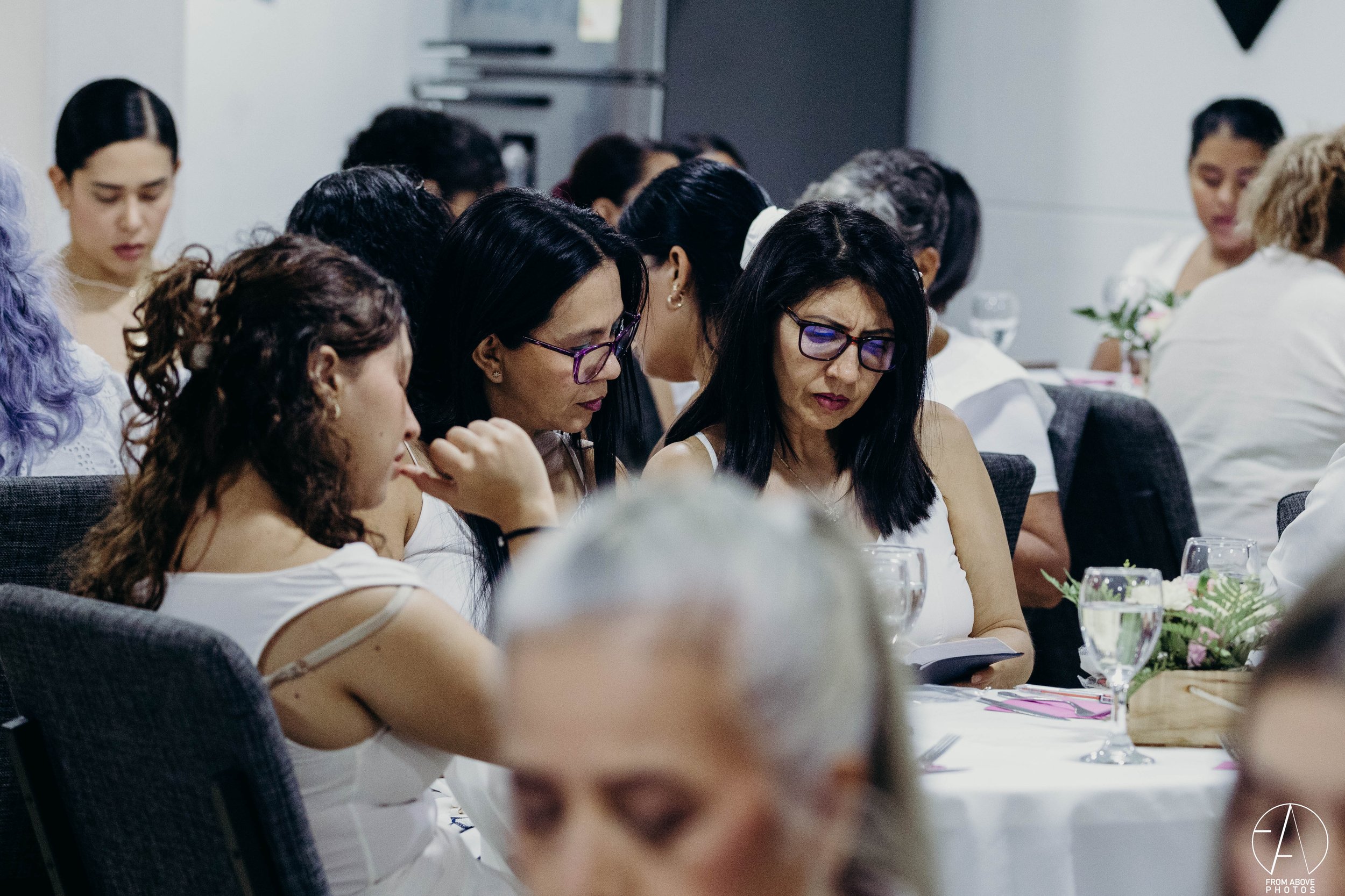 Grupo de mujeres en una mesa de reunión, algunas con lentes, en un ambiente de oficina, concentradas en lo que están leyendo o viendo en sus dispositivos, con decoraciones florales y cristalería en la mesa.