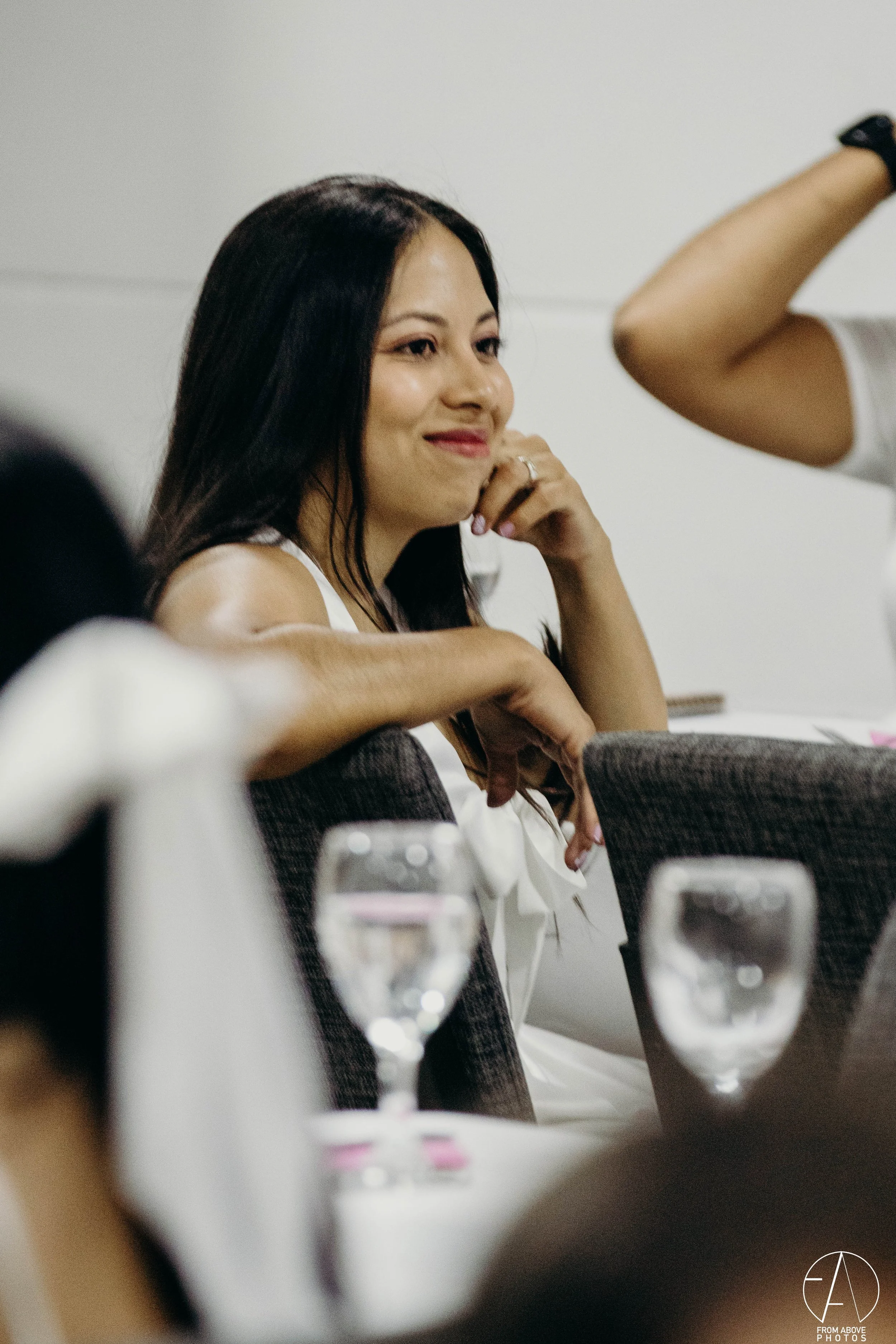 Mujer de cabello largo y oscuro, sonriendo, en una reunión o evento, rodeada de personas, con vasos y platos en una mesa.