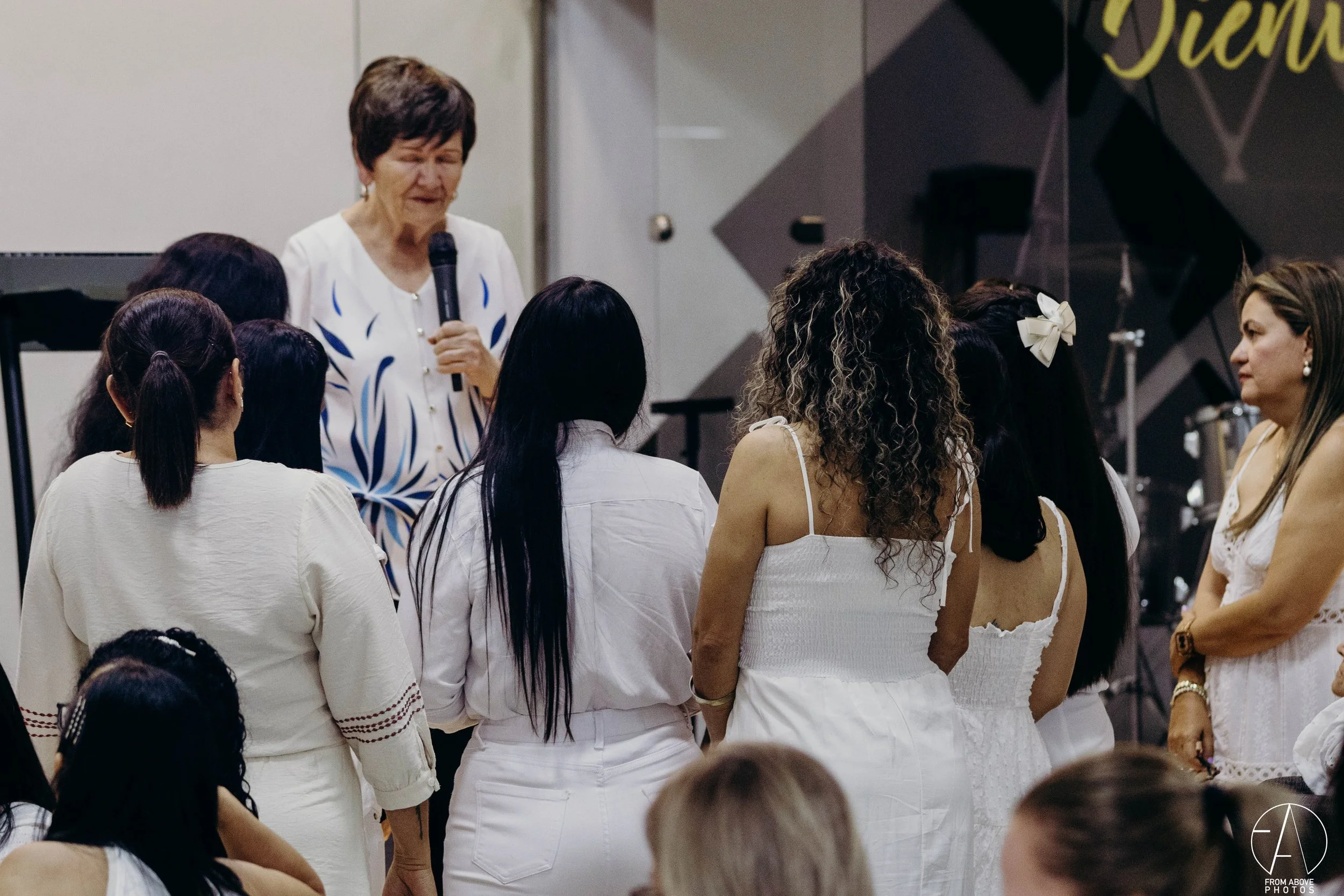 Mujer mayor con camiseta blanca y azul hablando con un micrófono a un grupo de mujeres jóvenes en un evento interior.