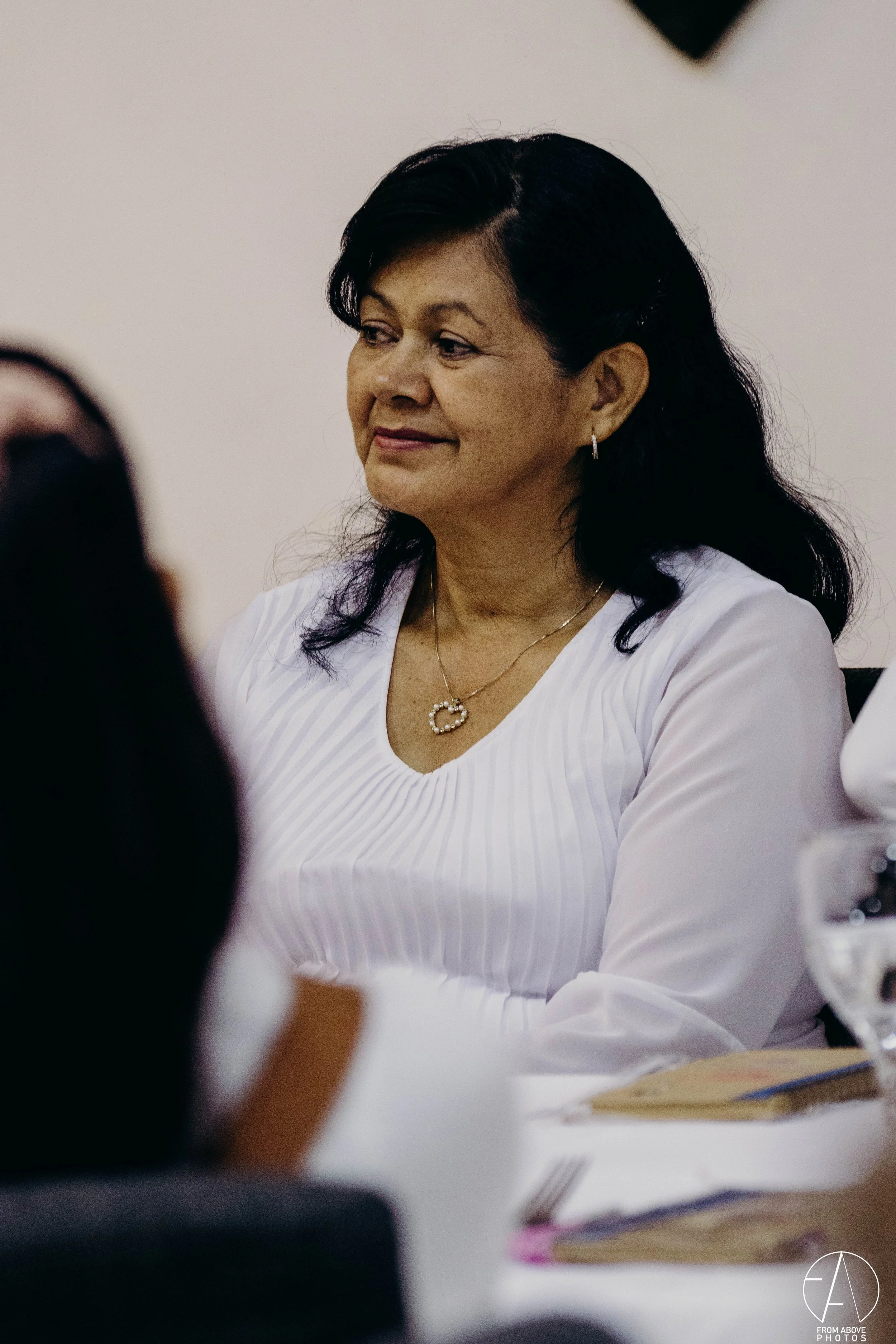 Mujer con cabello negro, short y blusa blanca, sentada en una mesa, en una reunión o evento formal, con expresión pensativa.