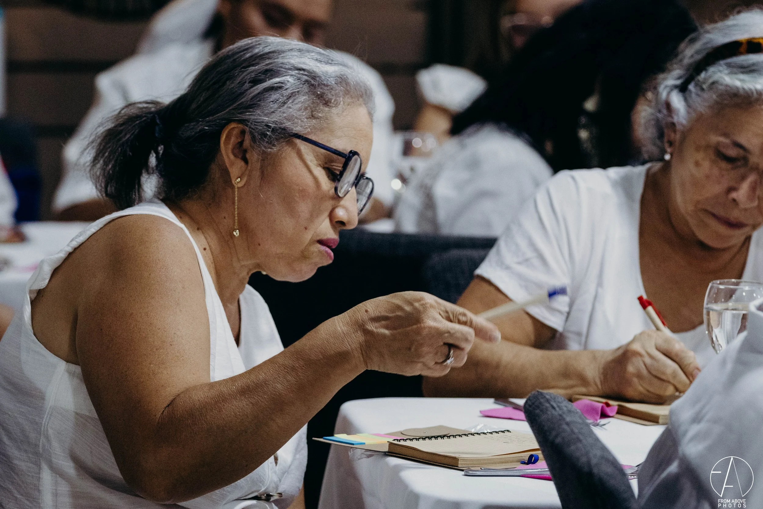 Mujeres mayores participando en una actividad de escritura en una mesa, con cuadernos y útiles de escritura, en un ambiente interior.