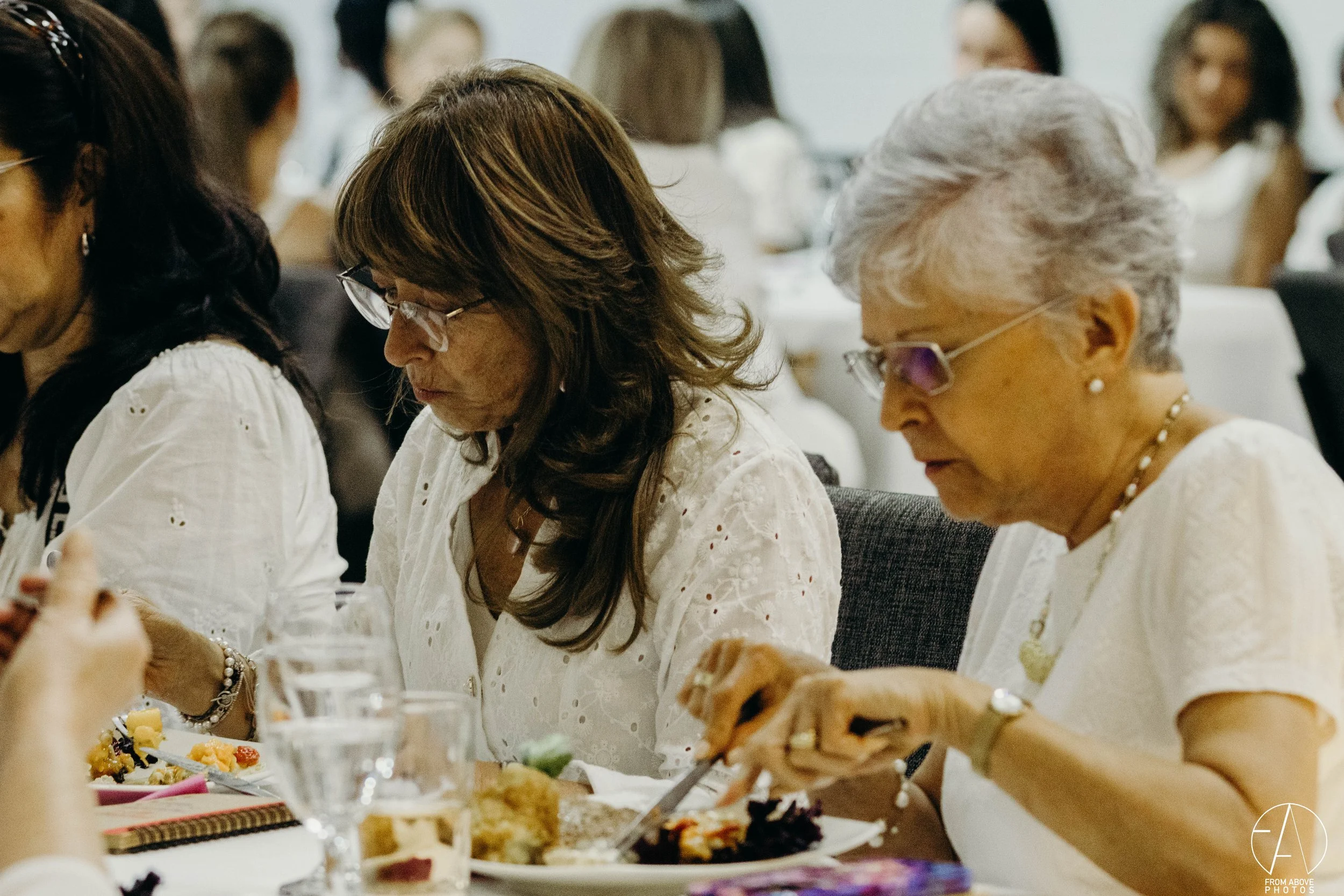 Mujeres mayores en una comida en una mesa, sirviéndose la comida y comiendo en un ambiente formal.