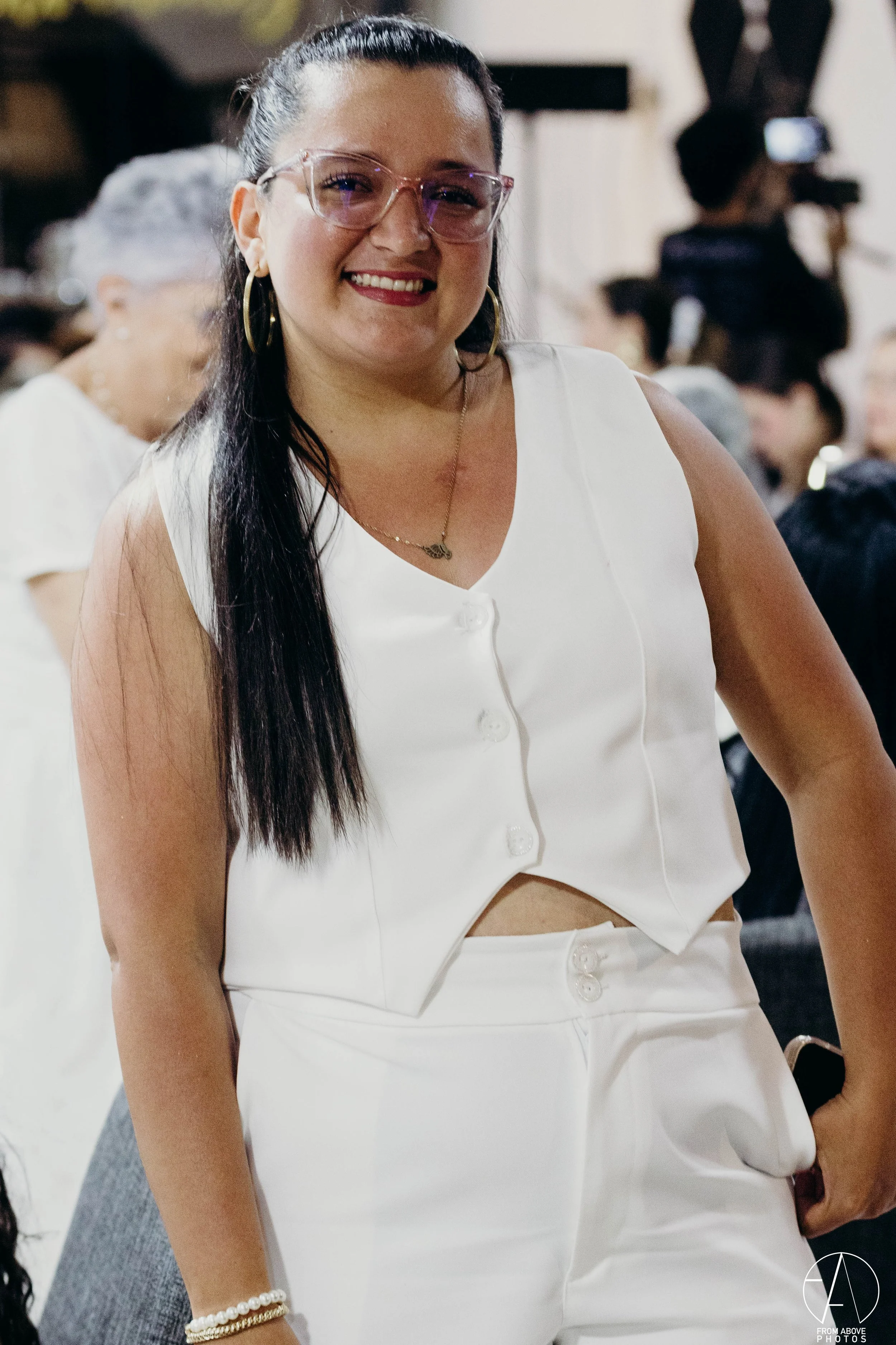 Joven mujer con gafas y pendientes grandes sonriendo en una reunión social, vestida con ropa blanca.