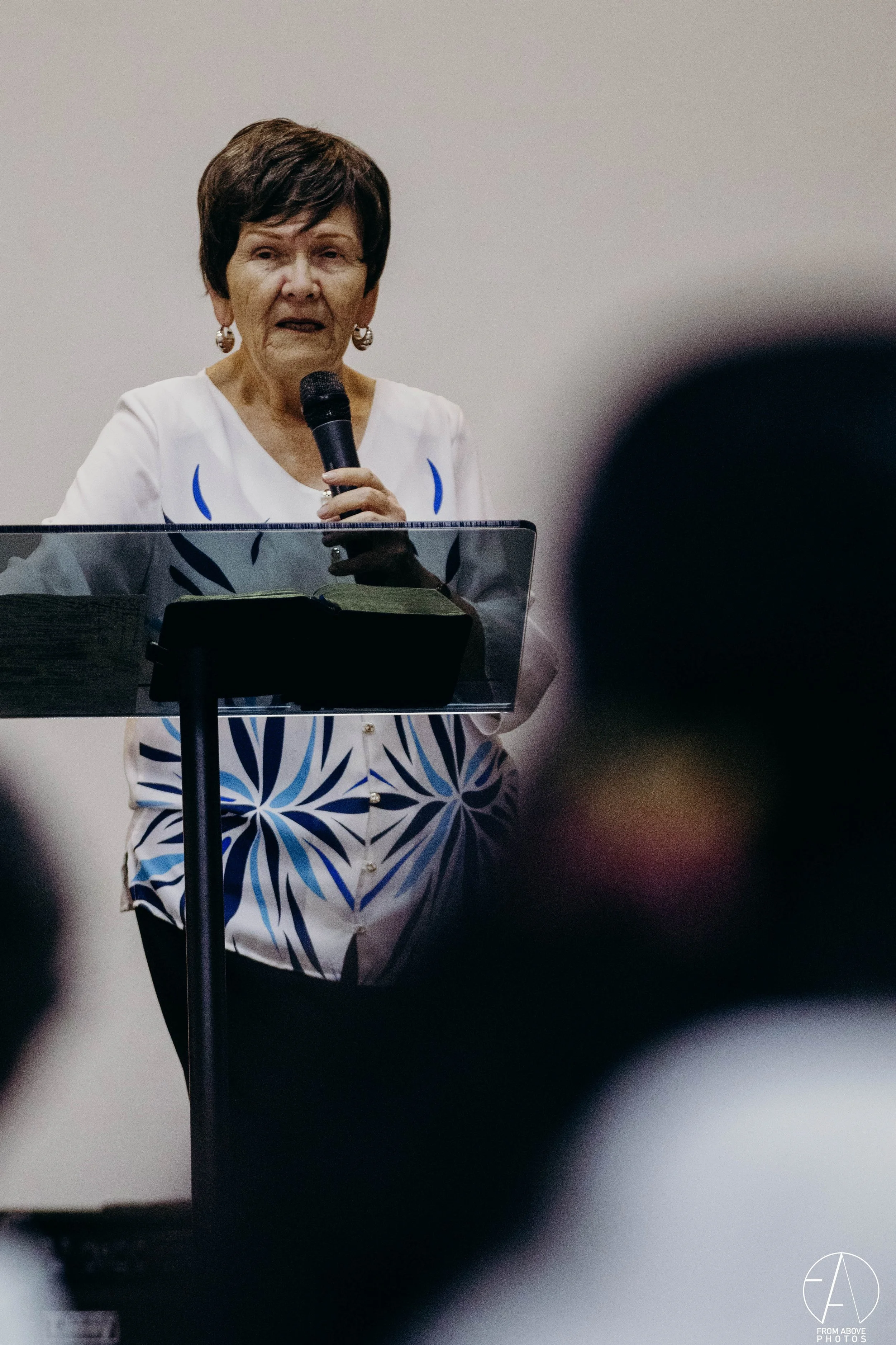 Una mujer mayor con cabello corto, vestida con blusa blanca con diseños azules, habla en un micrófono mientras lee de un libro en un atril