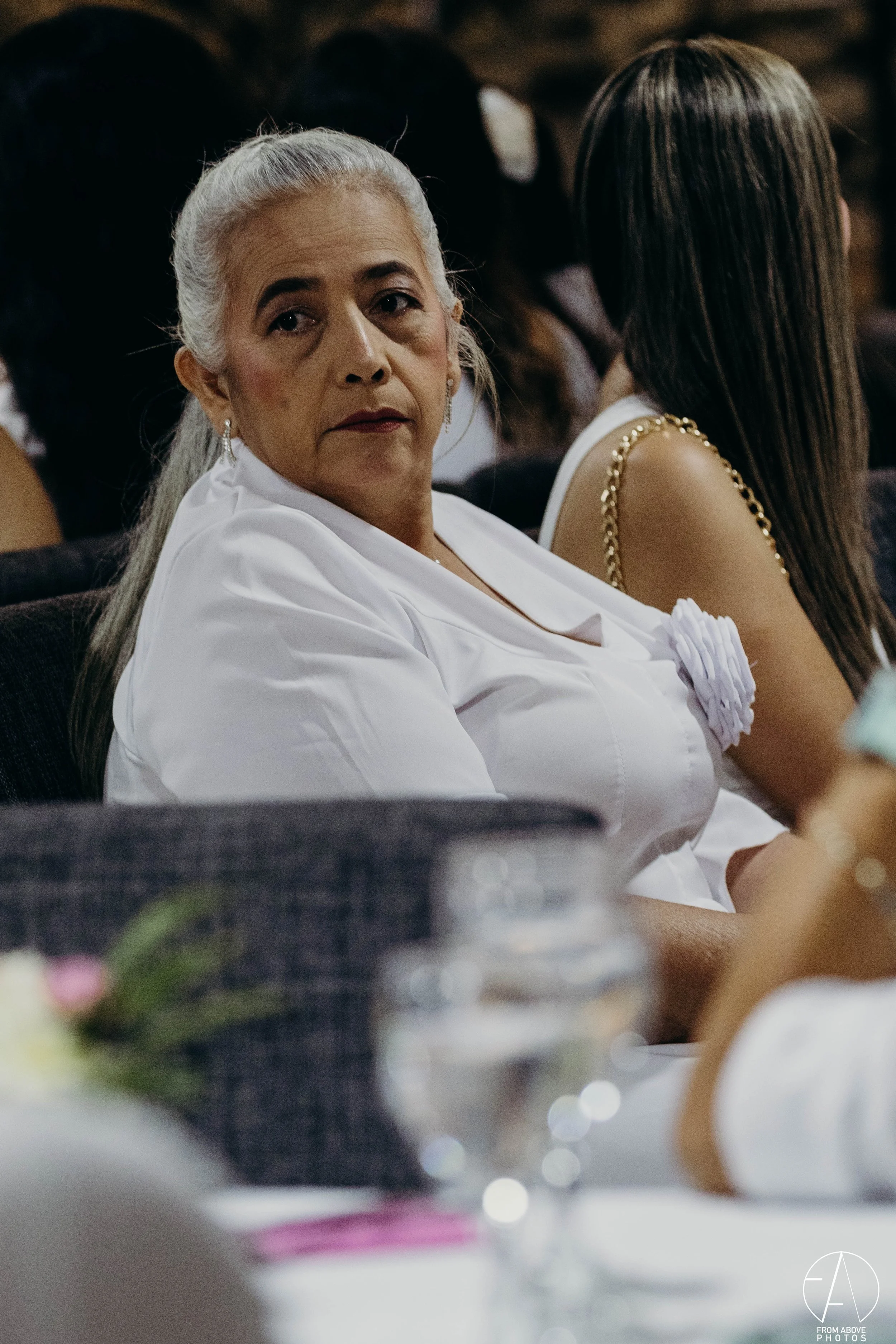 Mujer mayor con cabello gris peinado en cola de caballo, vestida con blusa blanca, sentada en un evento, con expresión seria, rodeada de otras personas también vestidas formalmente.