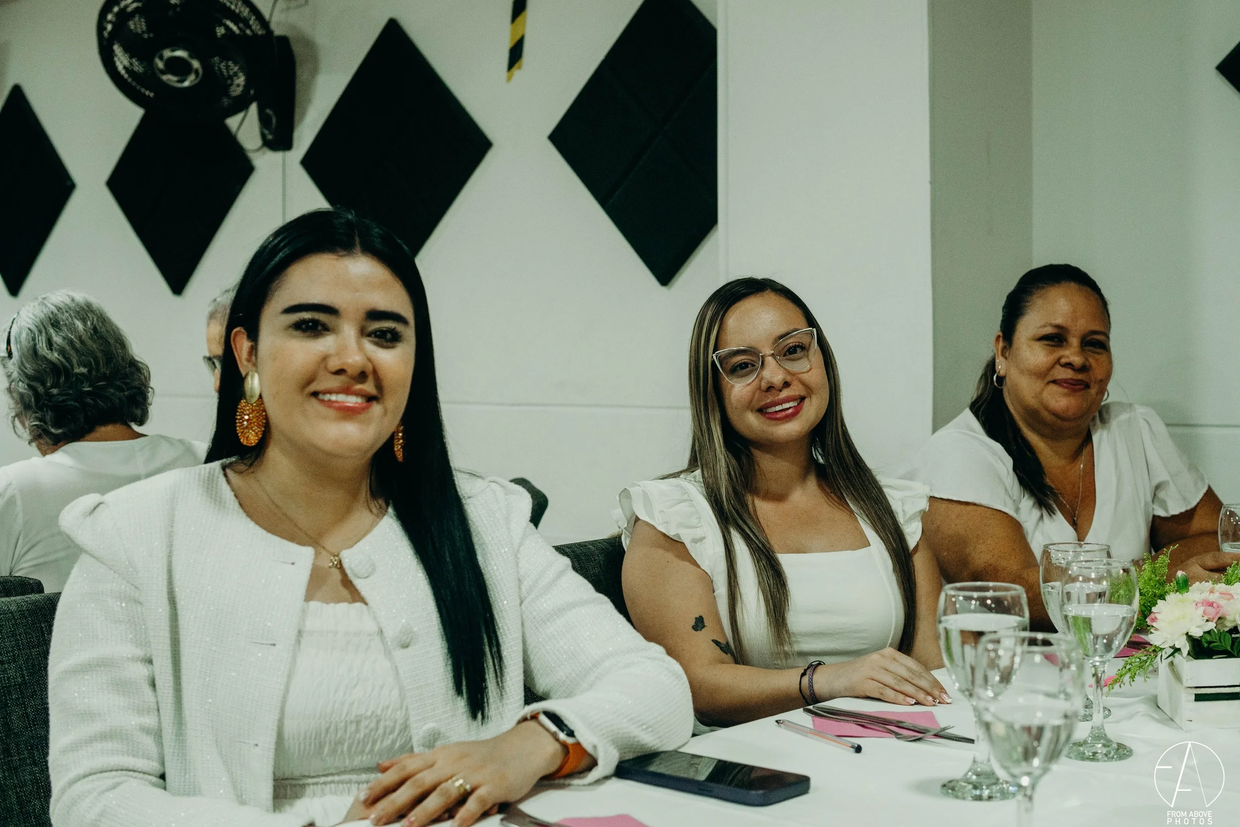 Tres mujeres sentadas en una mesa en un evento formal, sonriendo y con copas y flores sobre la mesa.