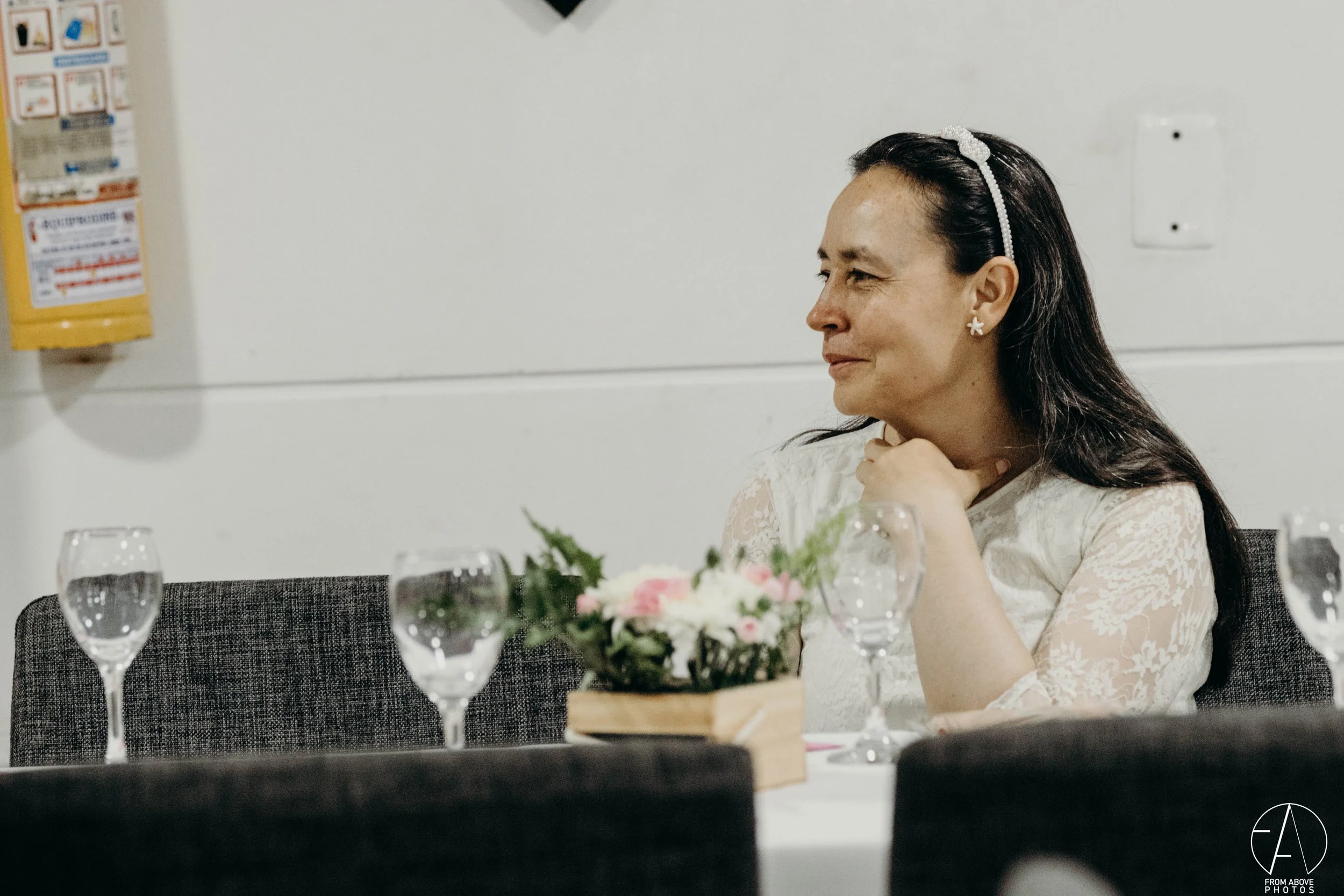 Mujer con cabello oscuro, bandas blancas y oreja de flor, sentada en una mesa con copas de agua y un ramo de flores, sonriendo y mirando hacia la derecha.