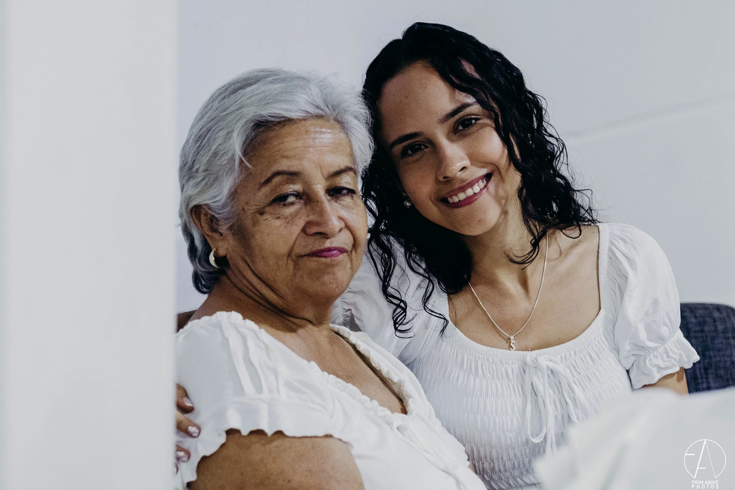 Dos mujeres, una anciana con cabello gris y otra joven con cabello rizado negro, sonrientes, en un ambiente interior con fondo blanco.