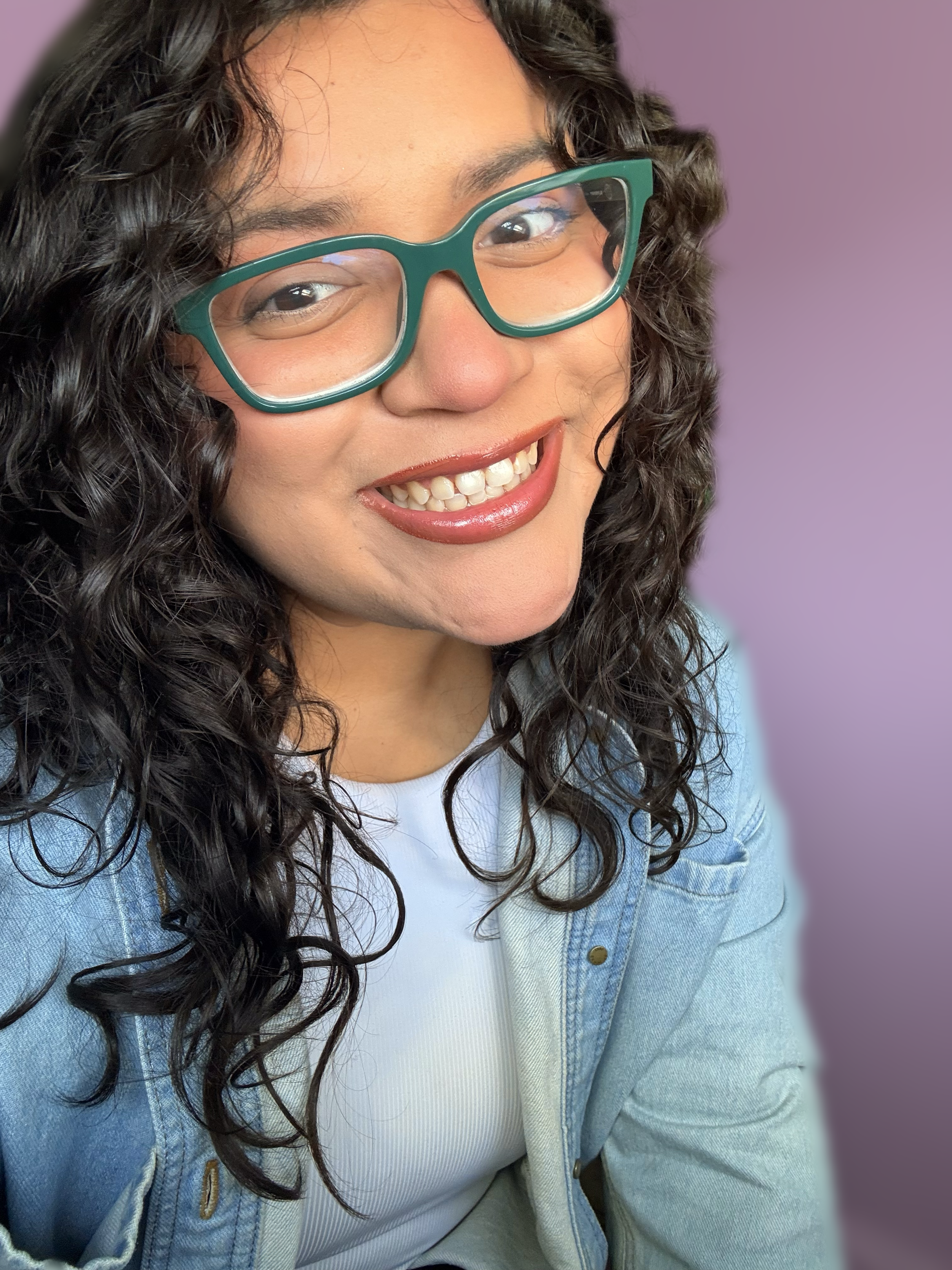 Close-up of a smiling woman with curly dark hair, wearing green glasses, a white shirt, and a light blue denim jacket, with a purple background. Founder of the design creative.
