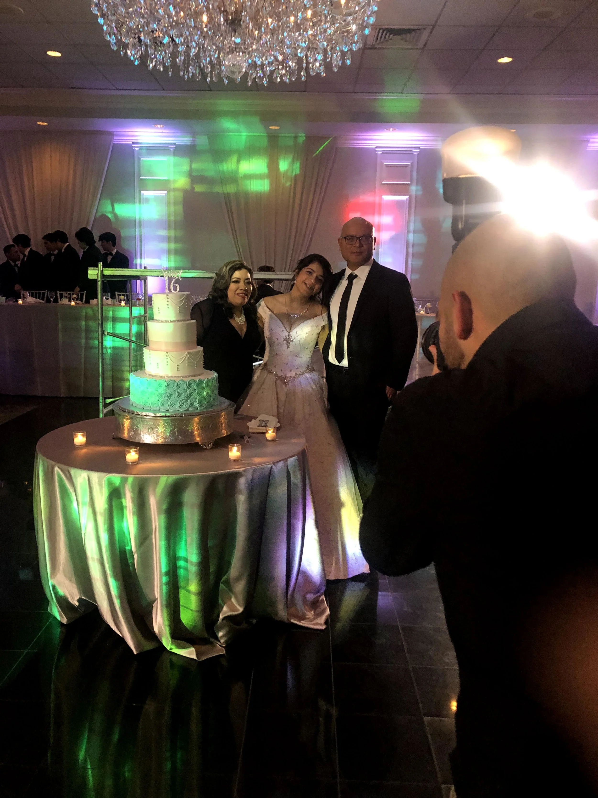 A wedding celebration with a bride and groom posing for a photo with a woman, in front of a tiered wedding cake on a round table, surrounded by candles, in a decorated reception hall with colorful lights and a chandelier.