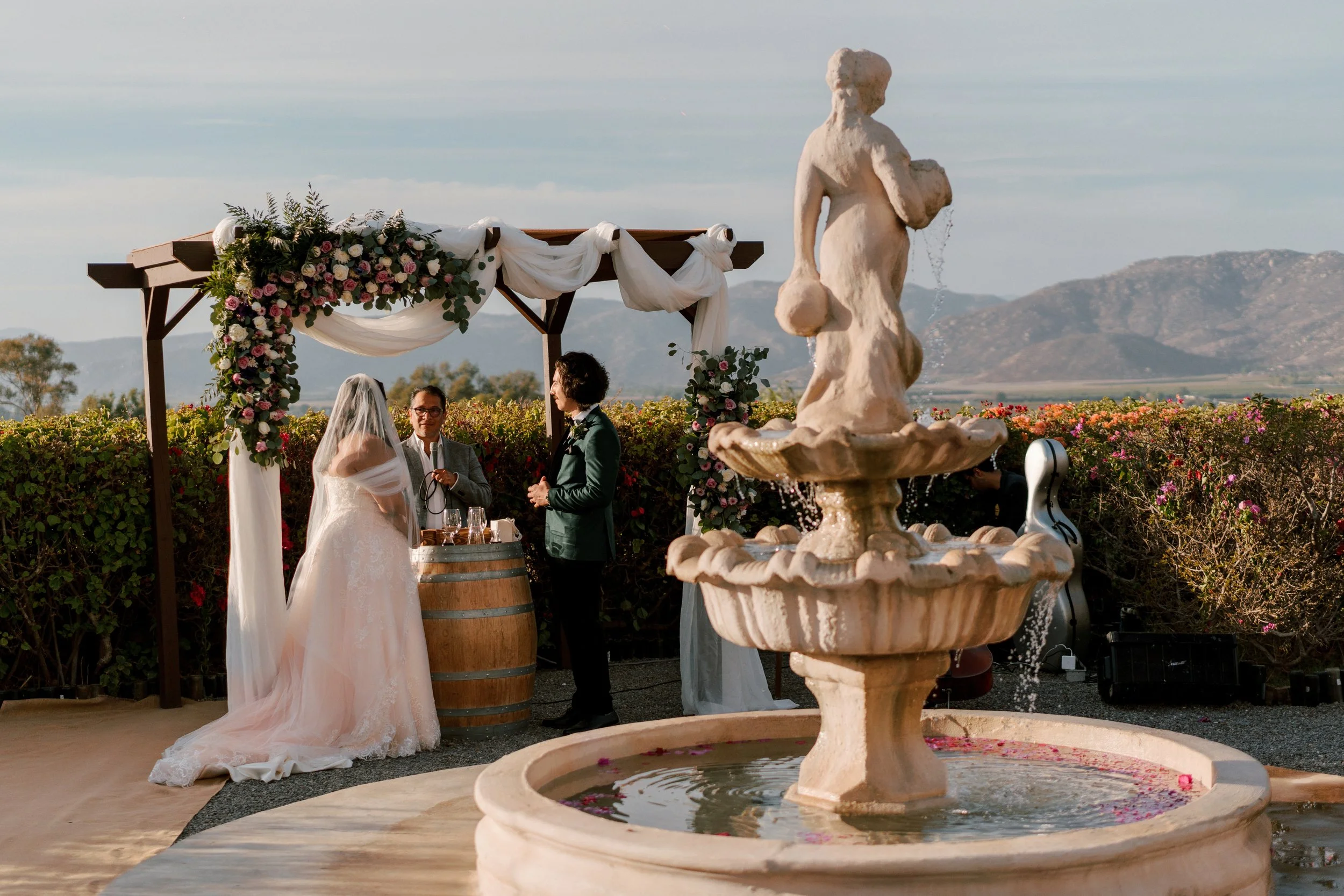 A wedding ceremony outdoors with a bride, groom, and officiant in front of a floral decorated arch. A large fountain with a cherub sculpture is prominently in the foreground, set against mountains and a scenic landscape.