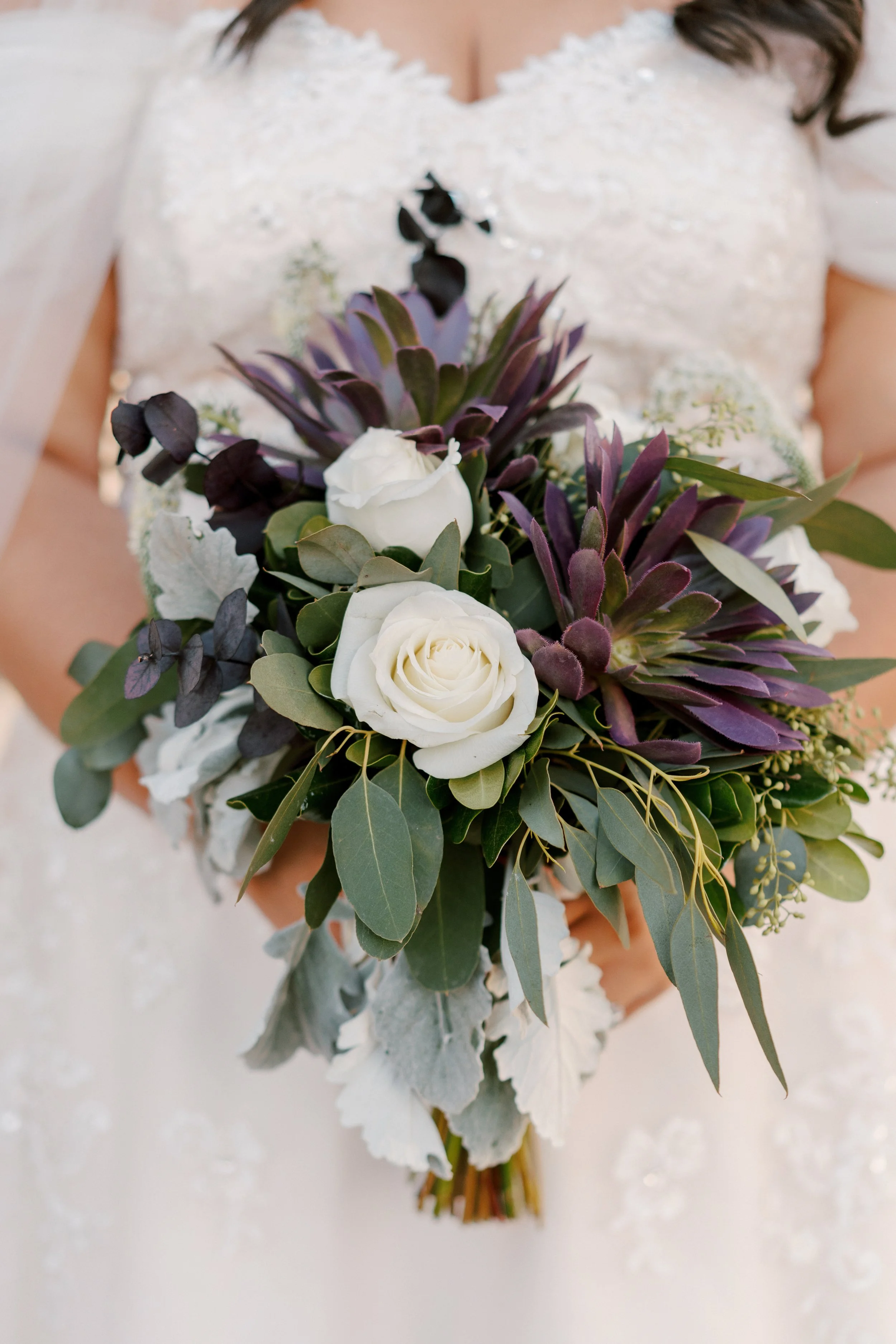 A bride holding a bouquet of white roses, purple succulents, and assorted greenery, wearing a wedding dress with lace details.