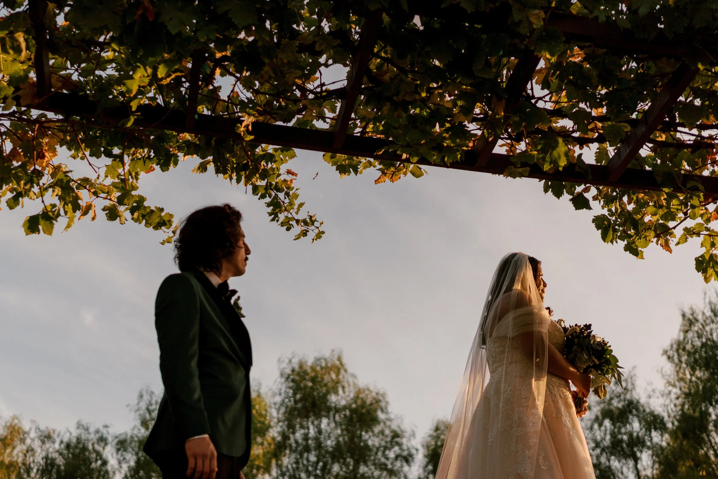 A bride in a wedding dress and veil holding a bouquet stands outdoors, facing a groom in a suit, during a wedding ceremony at sunset underneath a canopy of green leaves.