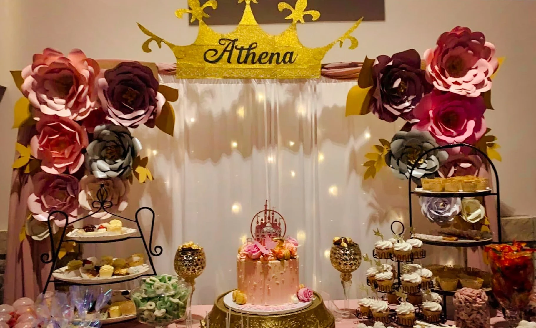 A decorated birthday party setup with a pink theme. There is a pink cake with flowers in the center, surrounded by baked goods and candies. A glittery gold crown with the name "Athena" at the top, large paper flowers in pink, purple, and white decorate the backdrop, with a sheer white curtain and fairy lights. Two cupcake stands display treats on either side.
