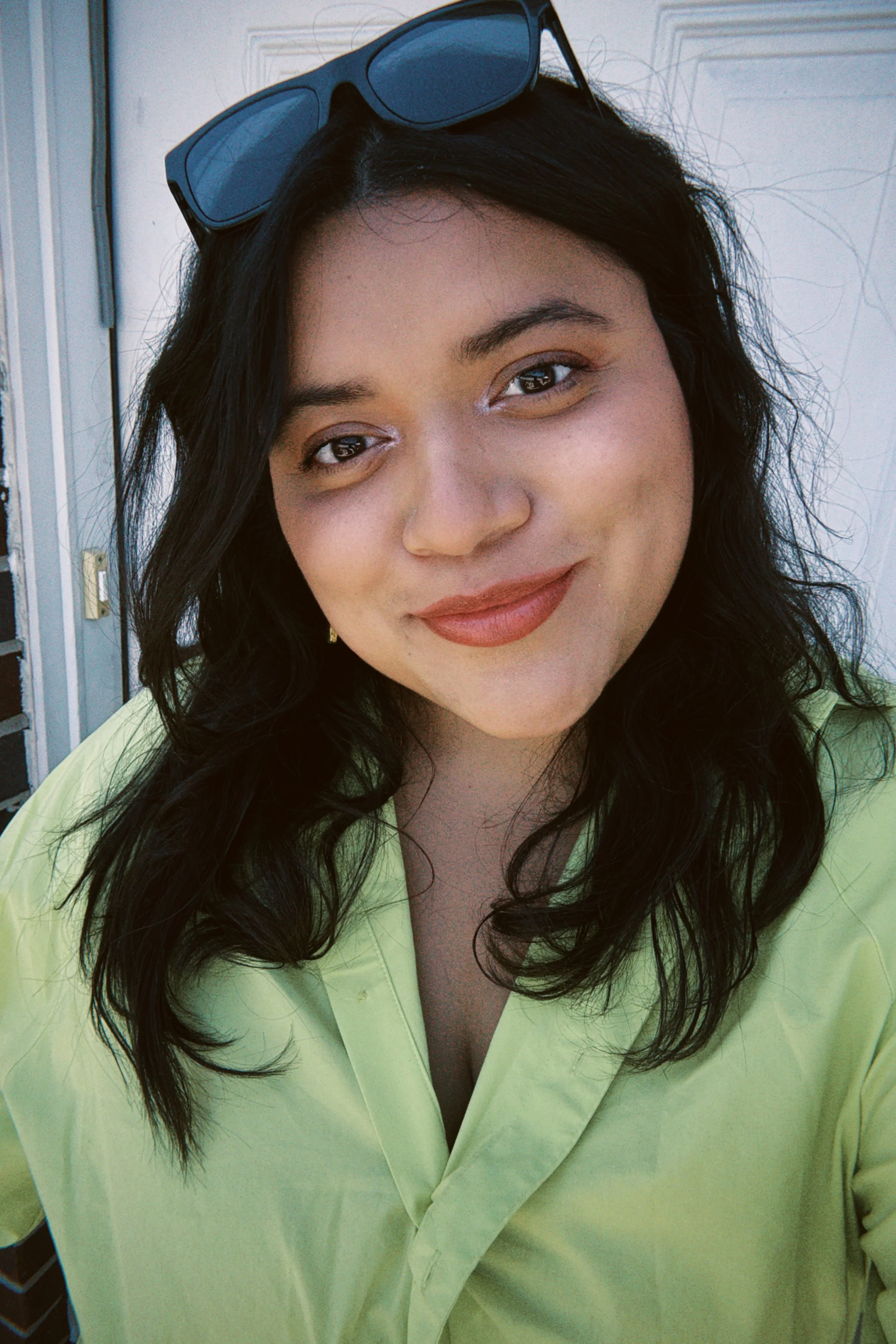 A young woman with dark wavy hair wearing sunglasses on her head, a yellow shirt, and makeup including lipstick, standing outdoors near a white wall.