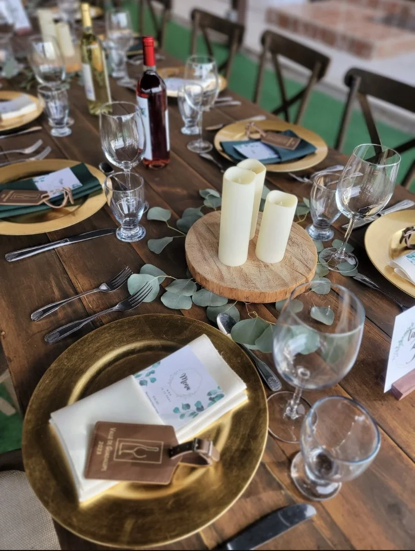 A formal dining table setup with gold charger plates, clear wine and water glasses, silverware, and place cards. A wooden centerpiece with three white candles is in the middle, surrounded by eucalyptus leaves. There are menus and tags on the table, and a bottle of red wine is also present.