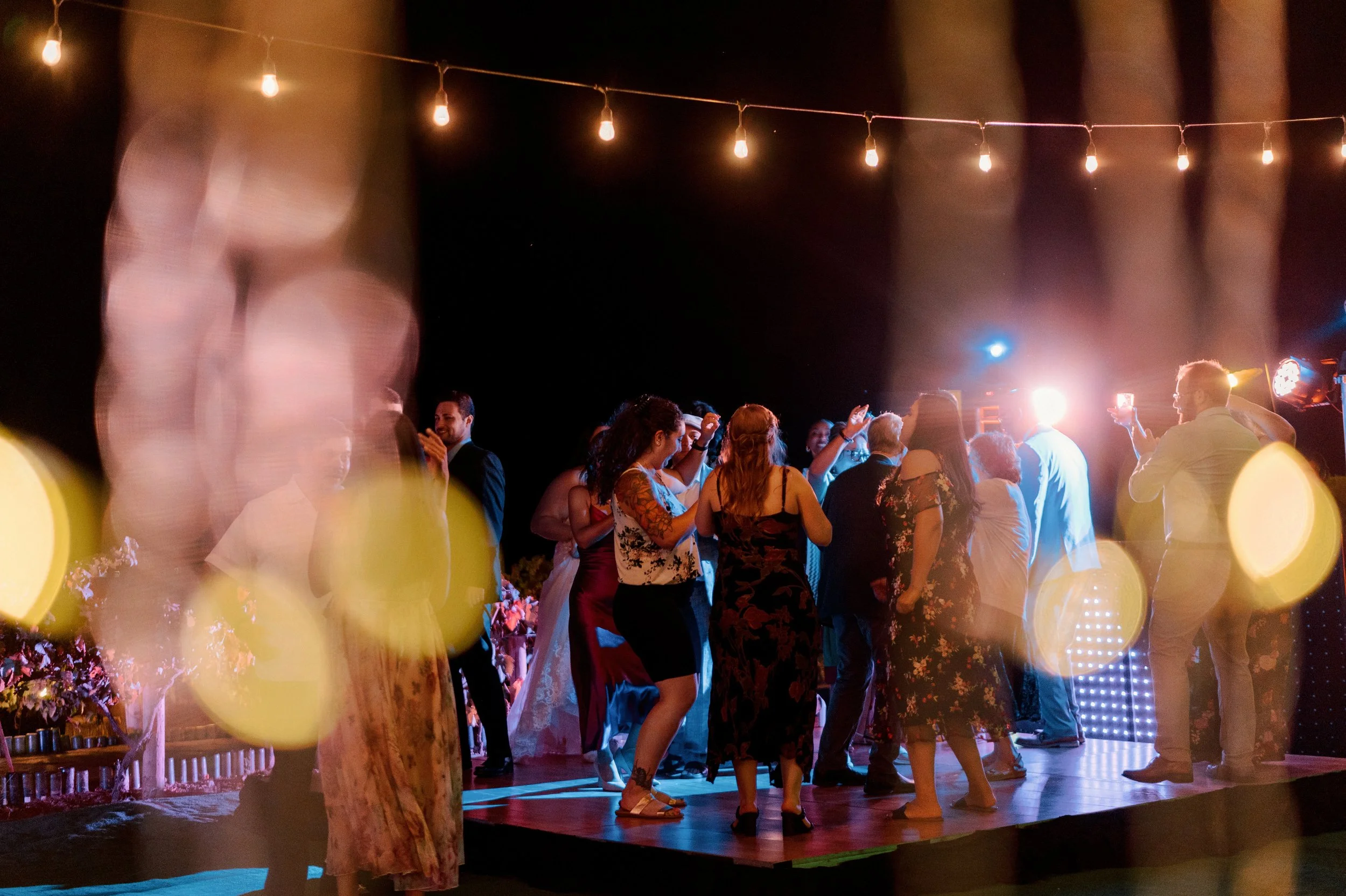 People dancing and socializing at an outdoor nighttime event under string lights, with colorful lighting effects and bokeh in the foreground.