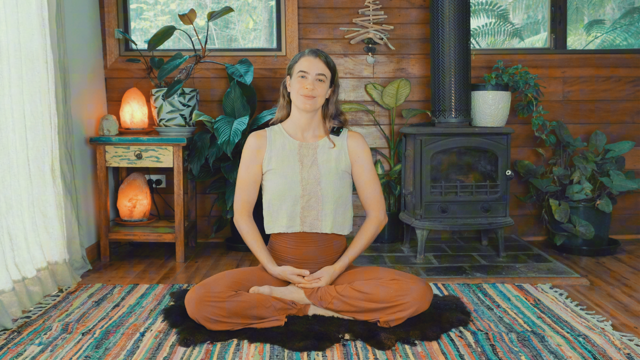 Woman sitting cross-legged on a colorful rug in a cozy, well-decorated room with plants, salt lamps, and a wood stove.