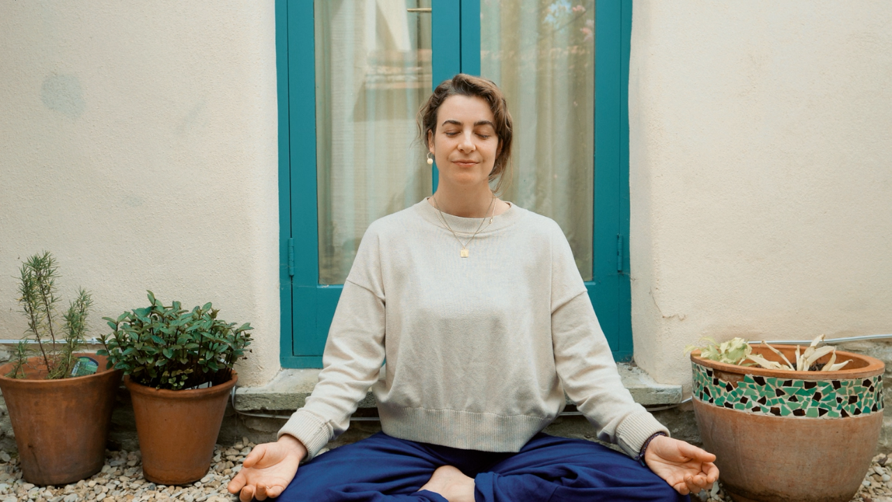 A woman practicing yoga outdoors, sitting cross-legged with eyes closed, in front of a turquoise door with potted plants on either side.