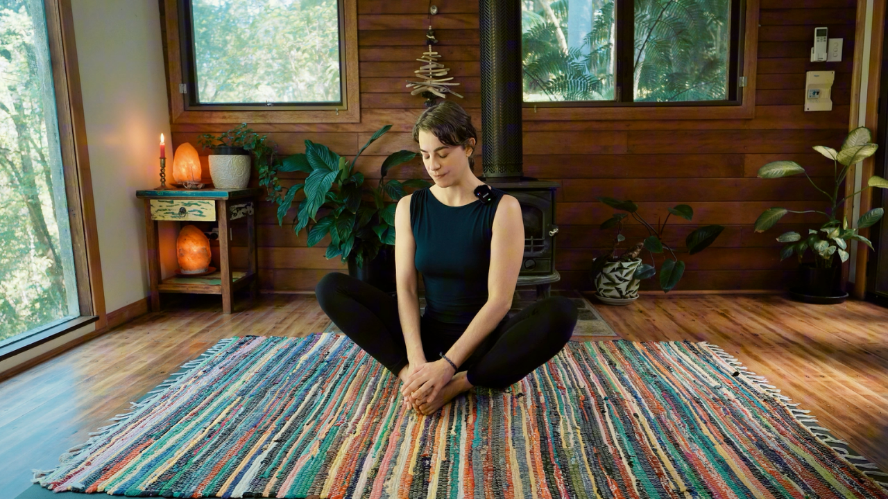 A woman sitting cross-legged on a colorful woven rug in a cozy, wooden interior space with plants and a wood stove.