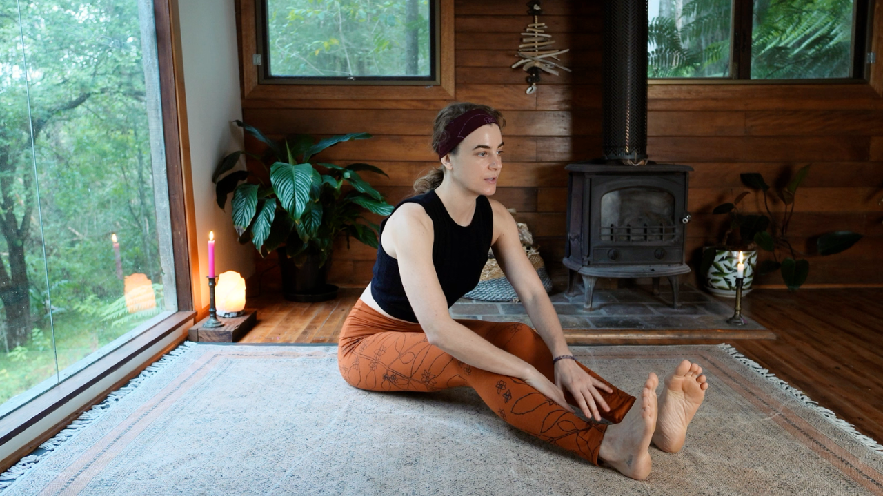 A woman practicing yoga indoors, sitting on a rug in a split position, reaching for her toes.