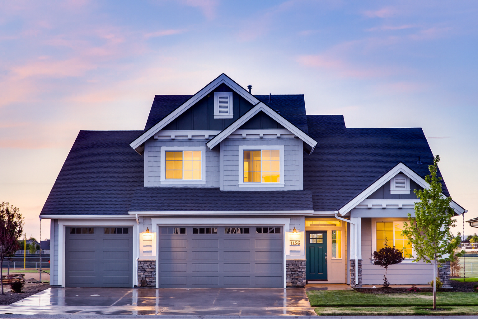 A two-story modern house with a dark roof, white and gray siding, illuminated windows, and a trim blue door, with a driveway, small trees, and a sunset sky in the background.
