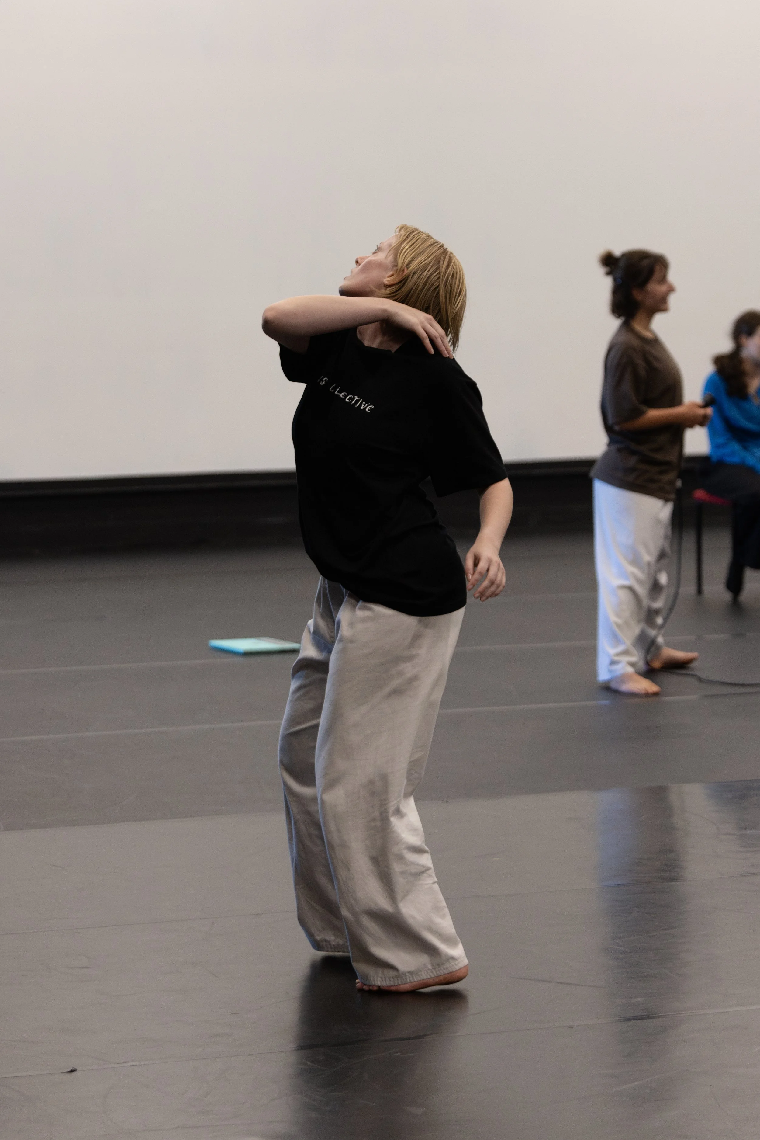 A woman with blonde hair practicing dance moves in a dance studio. She is wearing a black t-shirt and pants, standing barefoot on the black floor, with her right arm bent and across her neck, head tilted back, and eyes closed. In the background, two women are standing and looking to the side.