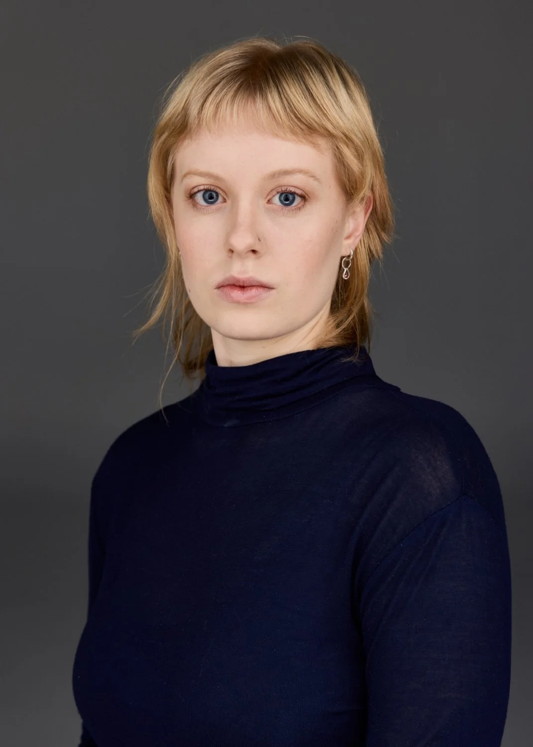 Portrait of a woman with short blonde hair, blue eyes, wearing a navy turtleneck and earrings, against a gray background.