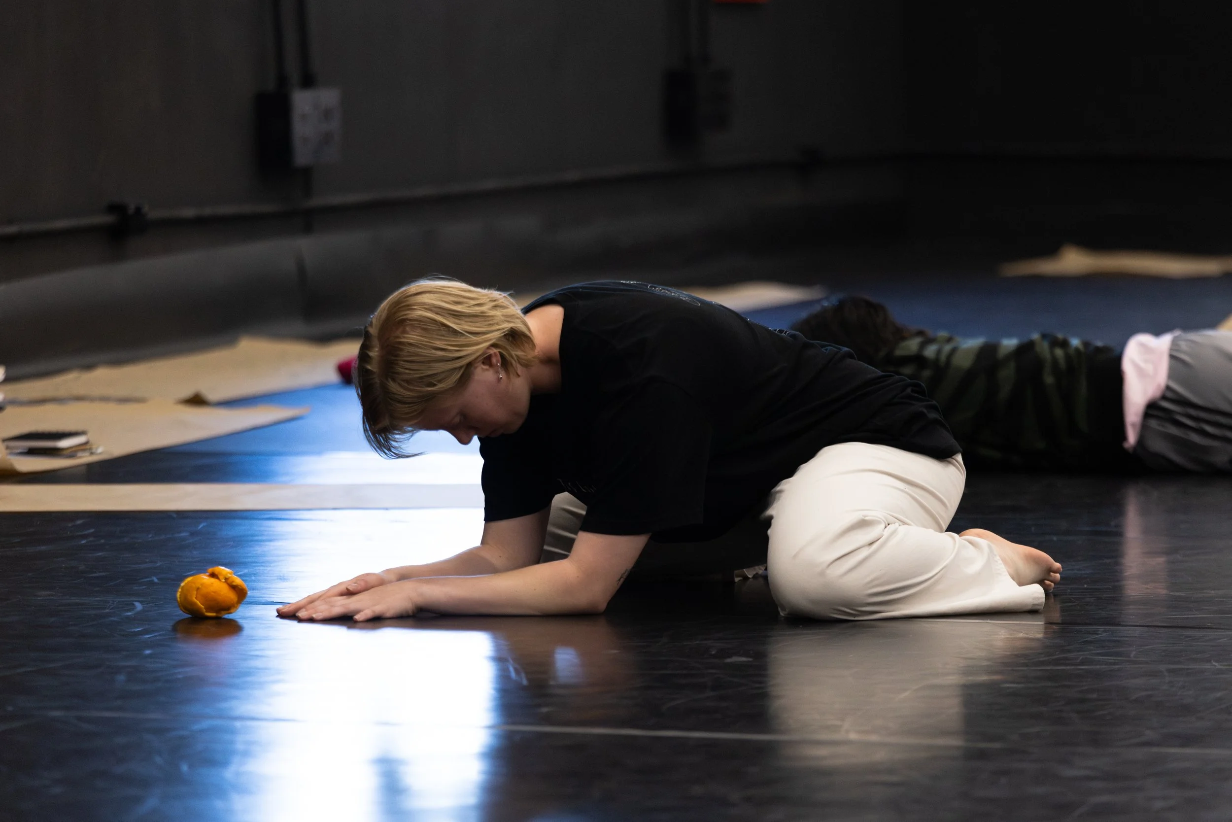 A woman kneeling on black studio floor, stretching forward with an orange in front of her, with other participants in background.