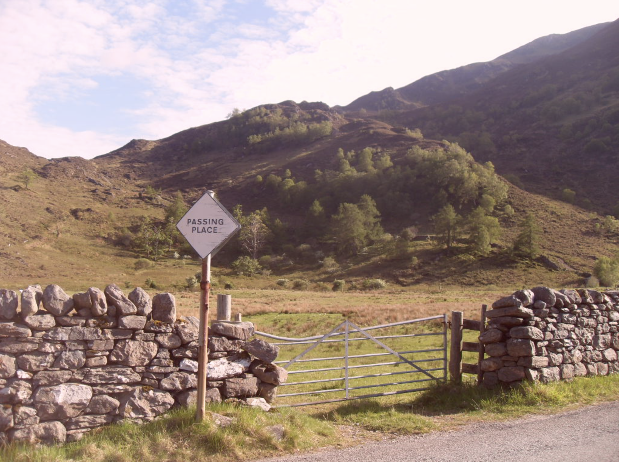 A rural stone wall with a metal gate, a sign that reads 'Passing Place,' and a scenic view of green hills and mountains in the background.