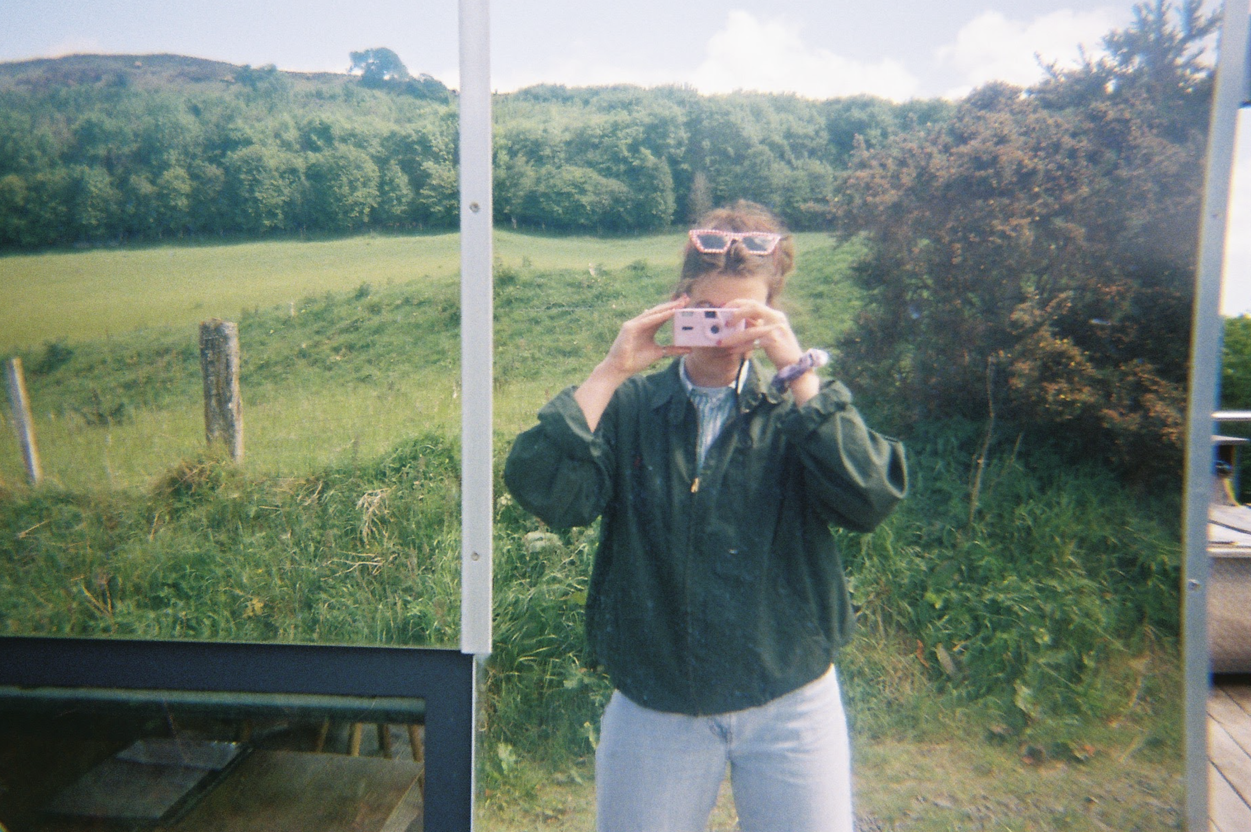 Person taking a photo in front of a mirror outdoors, with grassy fields and trees in the background.