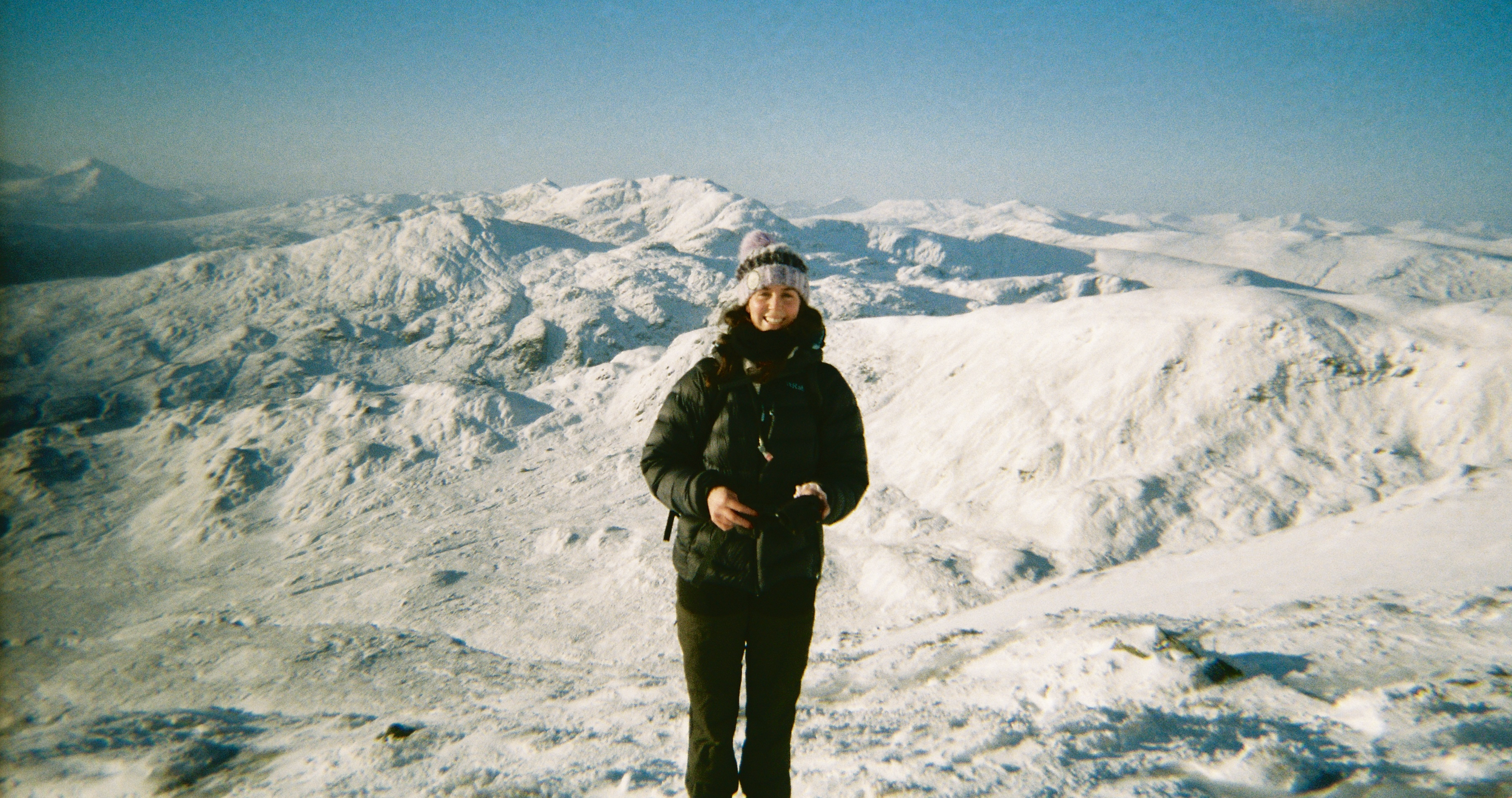 A woman dressed in winter clothing stands on a snow-covered mountain landscape with snowy peaks in the background under a clear blue sky.