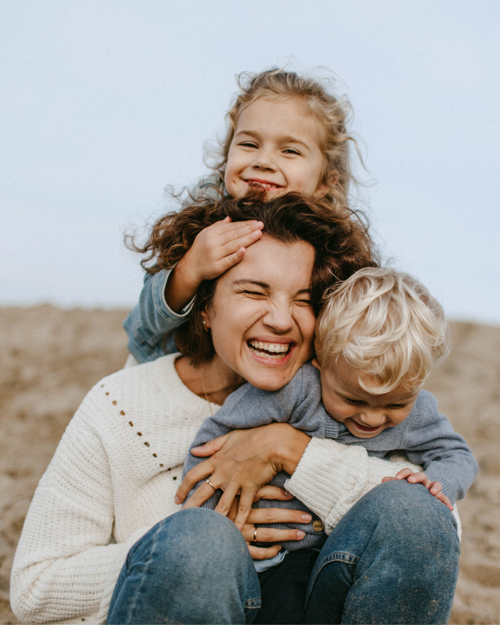 A woman with curly hair, wearing a white sweater, is smiling while holding two young children on her shoulders at the beach during daytime. One child has curly blond hair and is smiling, while the other with straight blond hair is resting on the woman's lap.