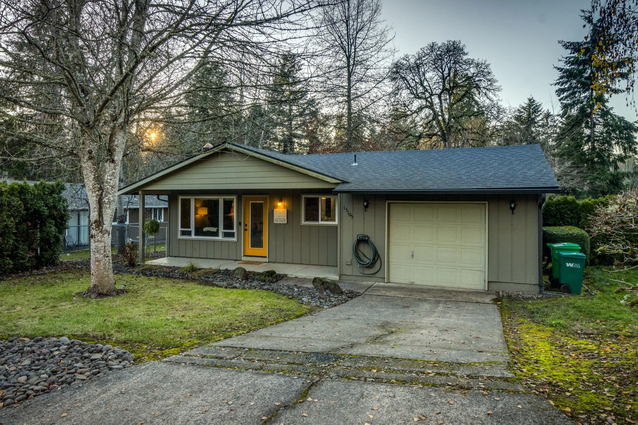 A single-story house with beige siding, a gray shingle roof, and front porch lights. There is a large tree in the front yard with bare branches. The driveway leads to an attached garage, with green recycling bins nearby. The sky is clear, and the sun is setting behind trees in the background.