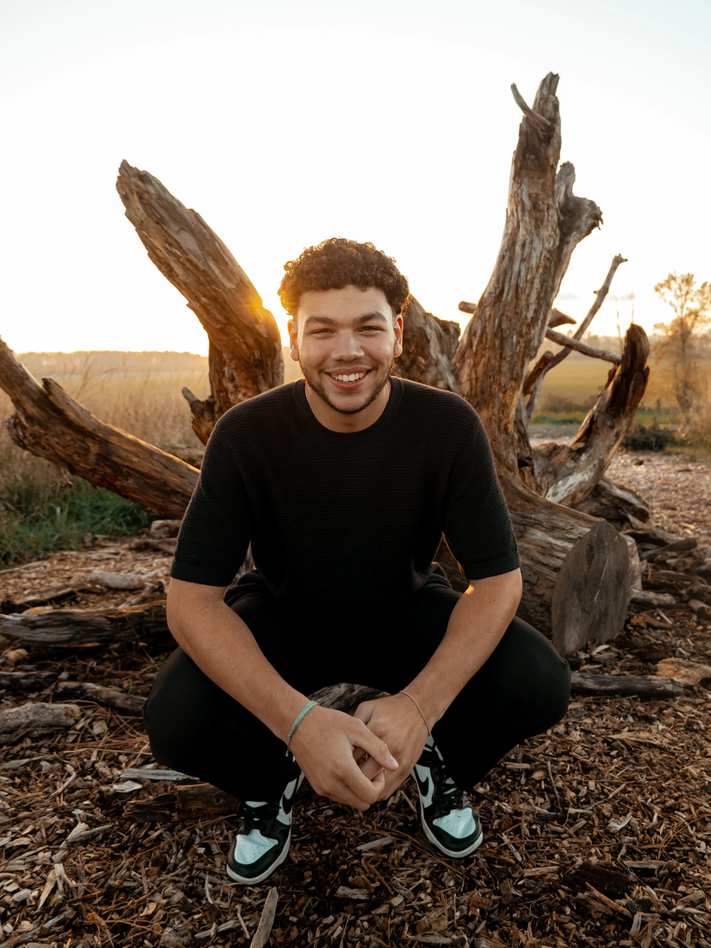 A young man with curly hair and a beard sitting cross-legged outdoors, smiling at the camera during sunset with a large, weathered tree trunk and a landscape in the background.
