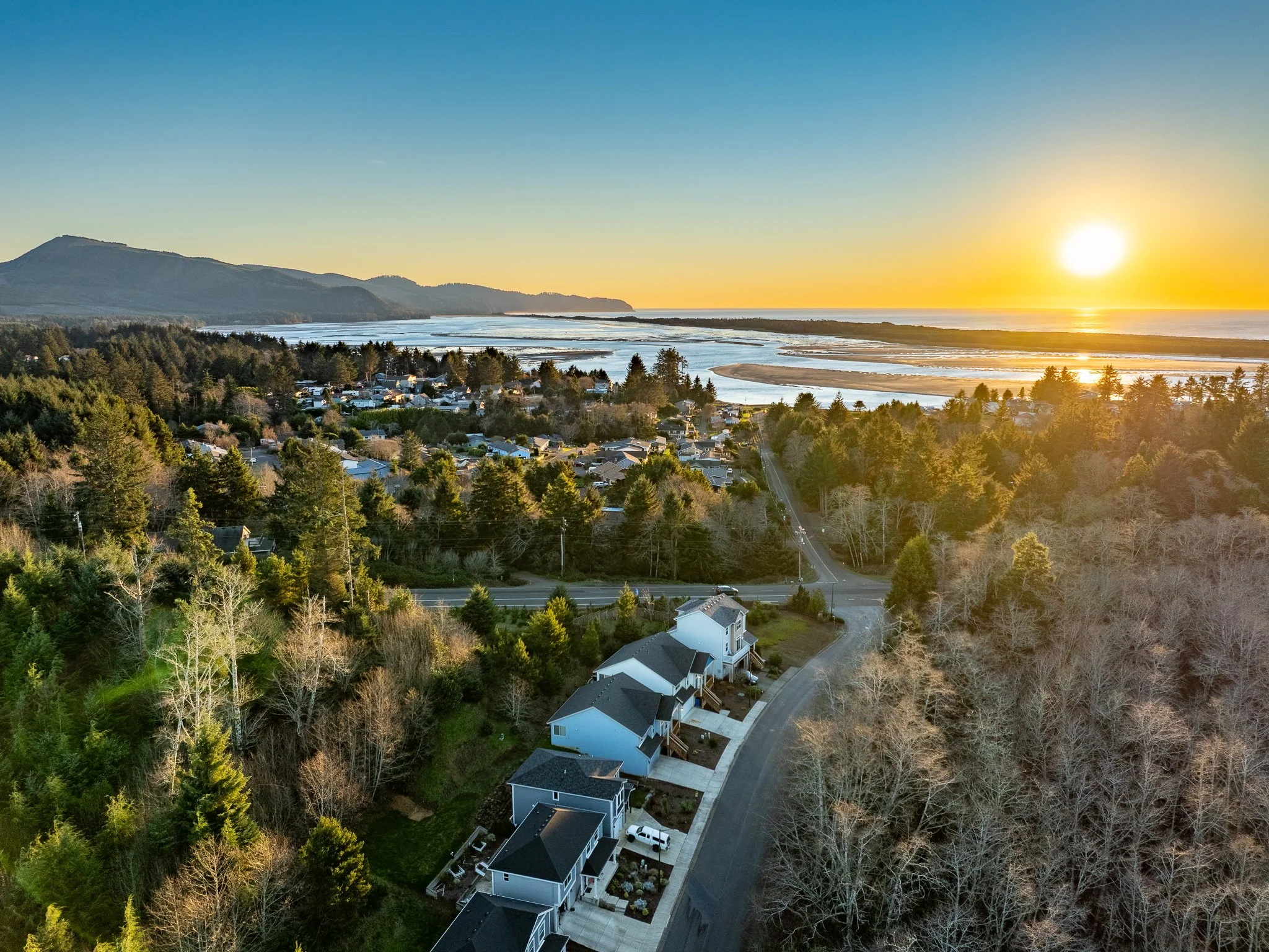A coastal town during sunset with houses along a winding road, dense green trees, ocean waves, and distant mountains under a clear sky.