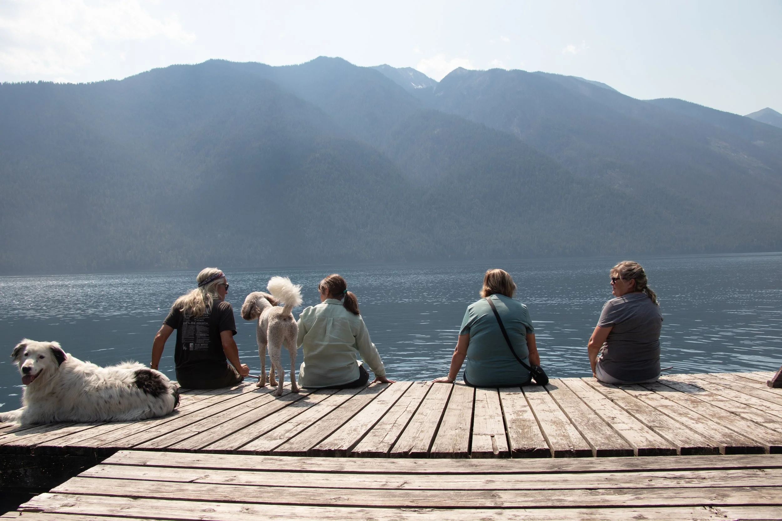 Four women and two dogs sitting on a wooden dock by a lake with mountain range in the background.