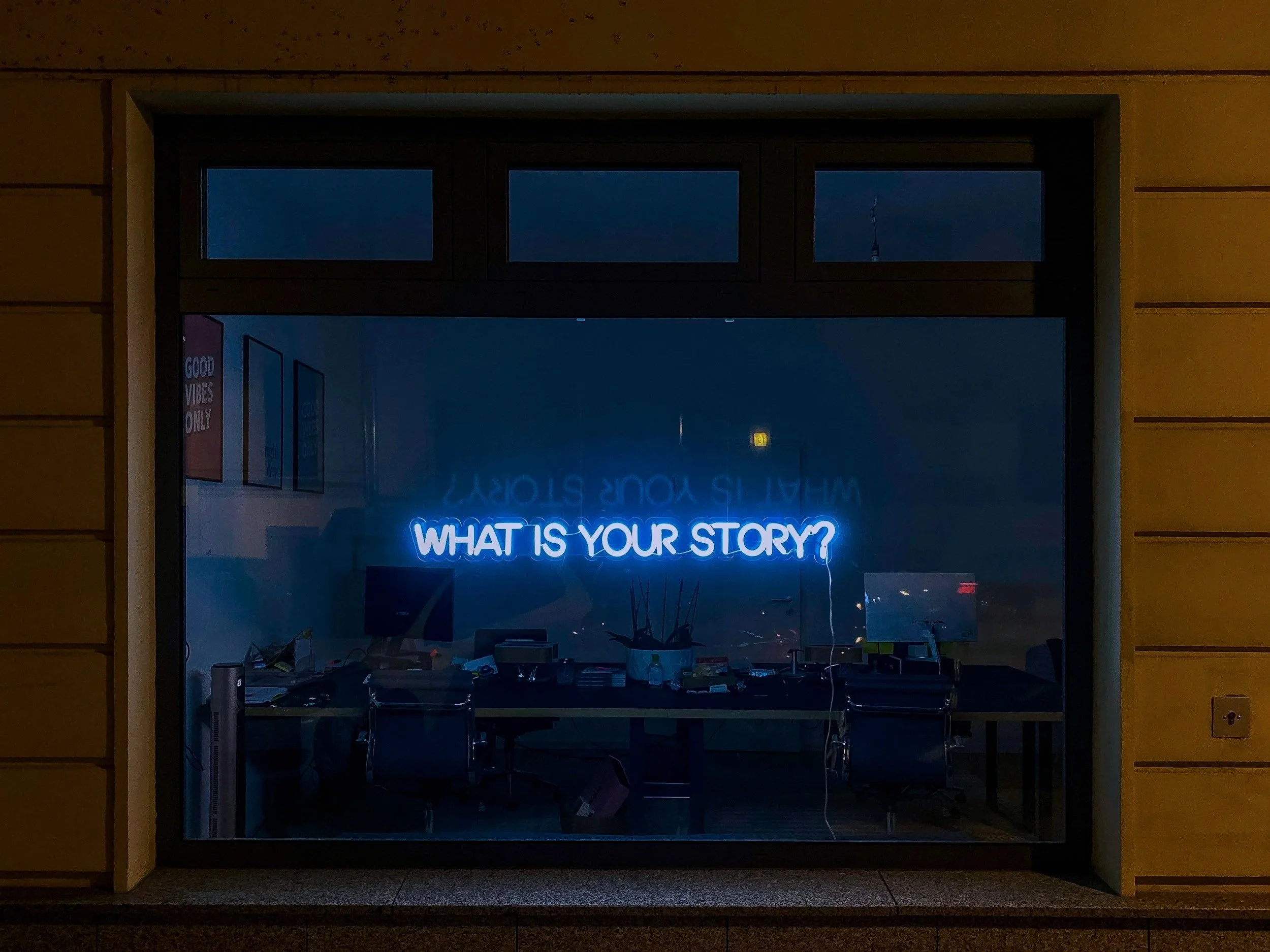 Neon sign in a window that reads 'WHAT IS YOUR STORY?' with office desks and chairs inside, and a sign on the wall that says 'GOOD VIBES ONLY'.