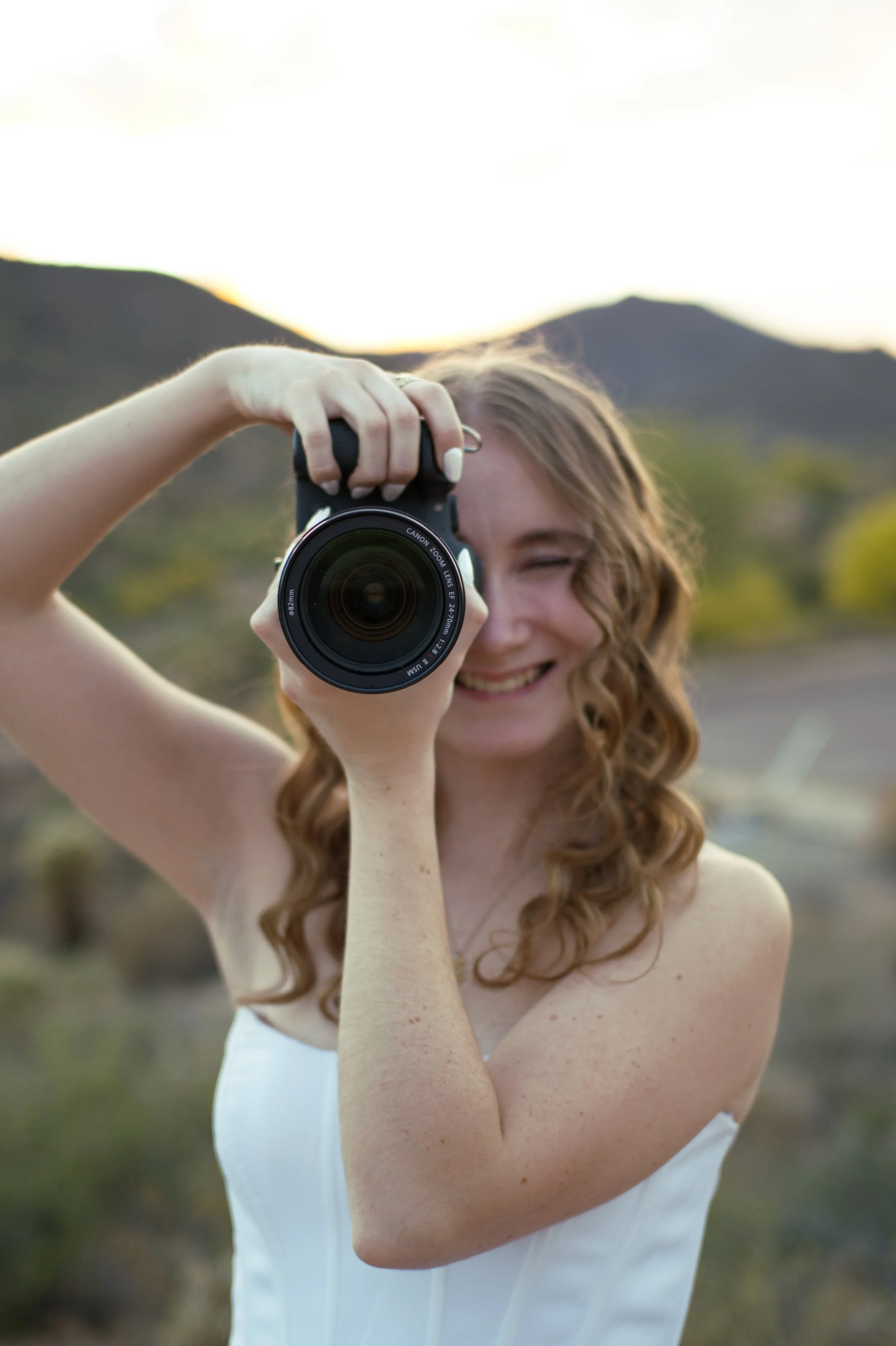 Candid close-up portrait of Amanda laughing while holding a camera up to take a photo, at Barrett Lake, Arizona, against desert mountains at golden hour.