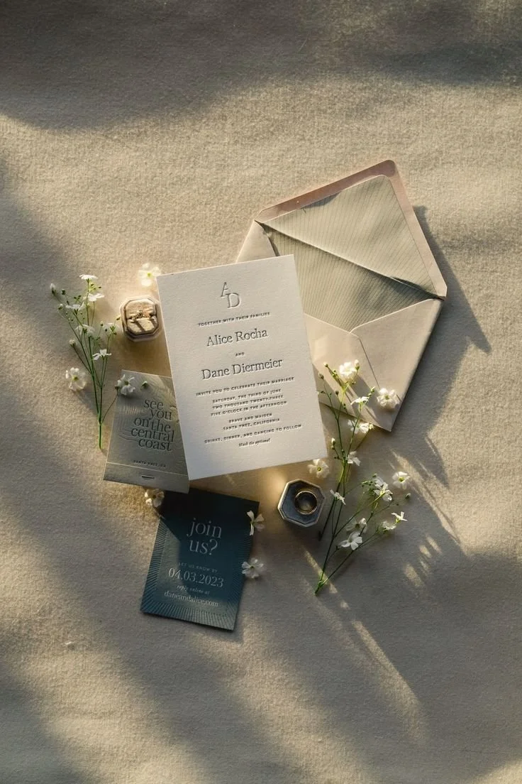 Wedding invitation and event details surrounded by small white flowers, a ring box, a small hexagon-shaped ring holder, and an open envelope on a textured beige surface, bathed in warm sunlight.