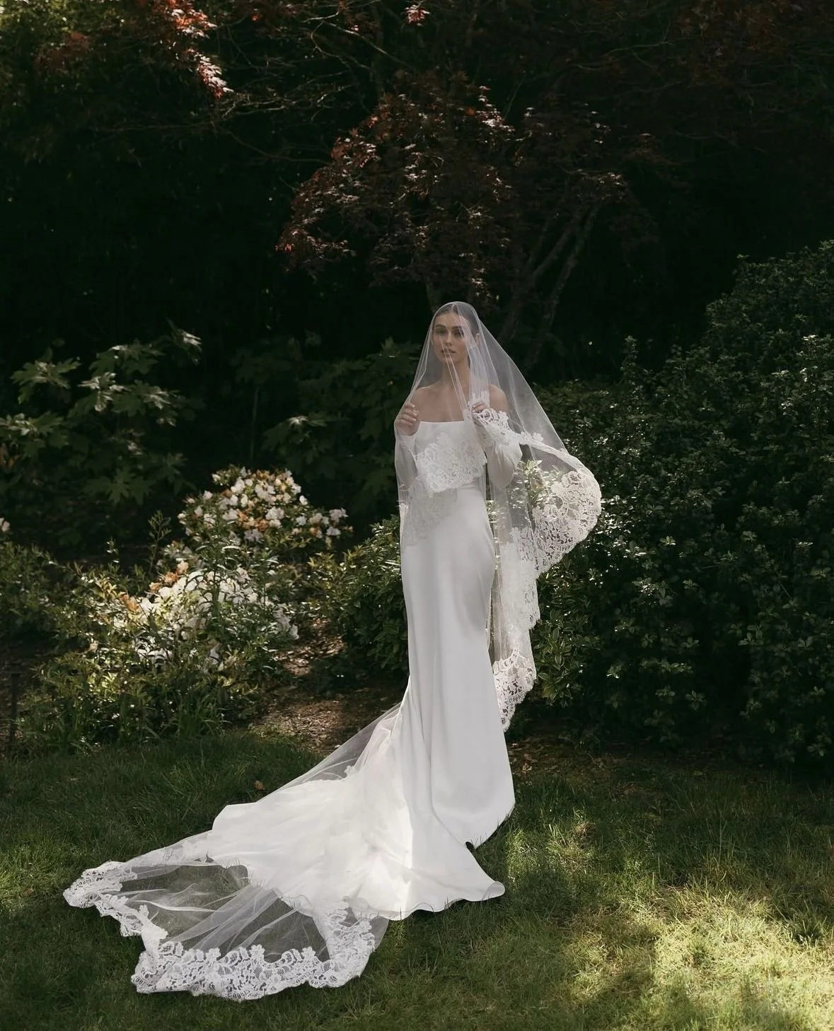 A bride in a white gown with a long train and lace veil standing outdoors among greenery and flowers.