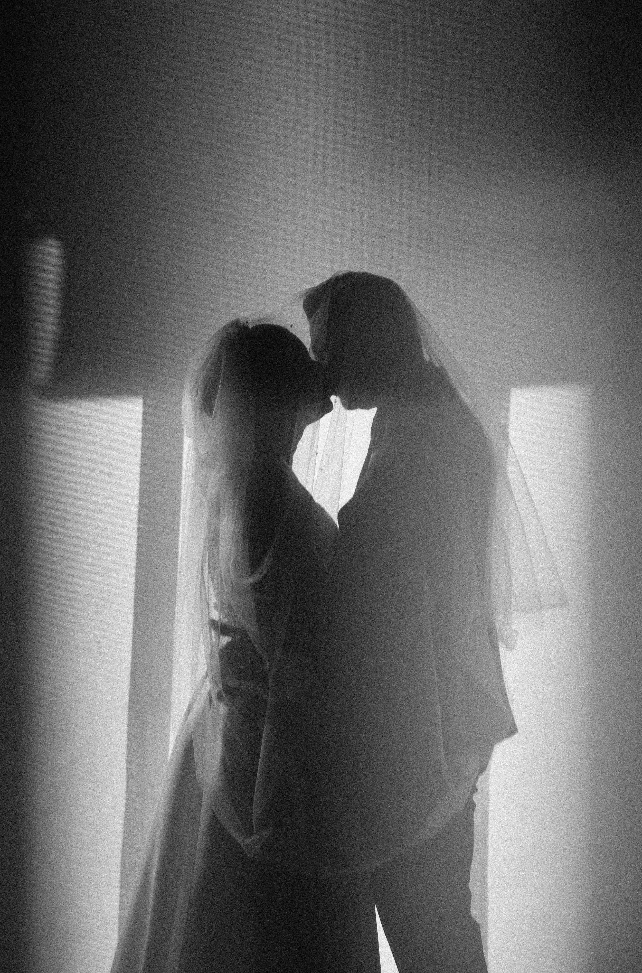 Black and white photograph of a bride and groom in a close embrace, wearing wedding veils, with their profiles touching, backlit to create silhouettes.