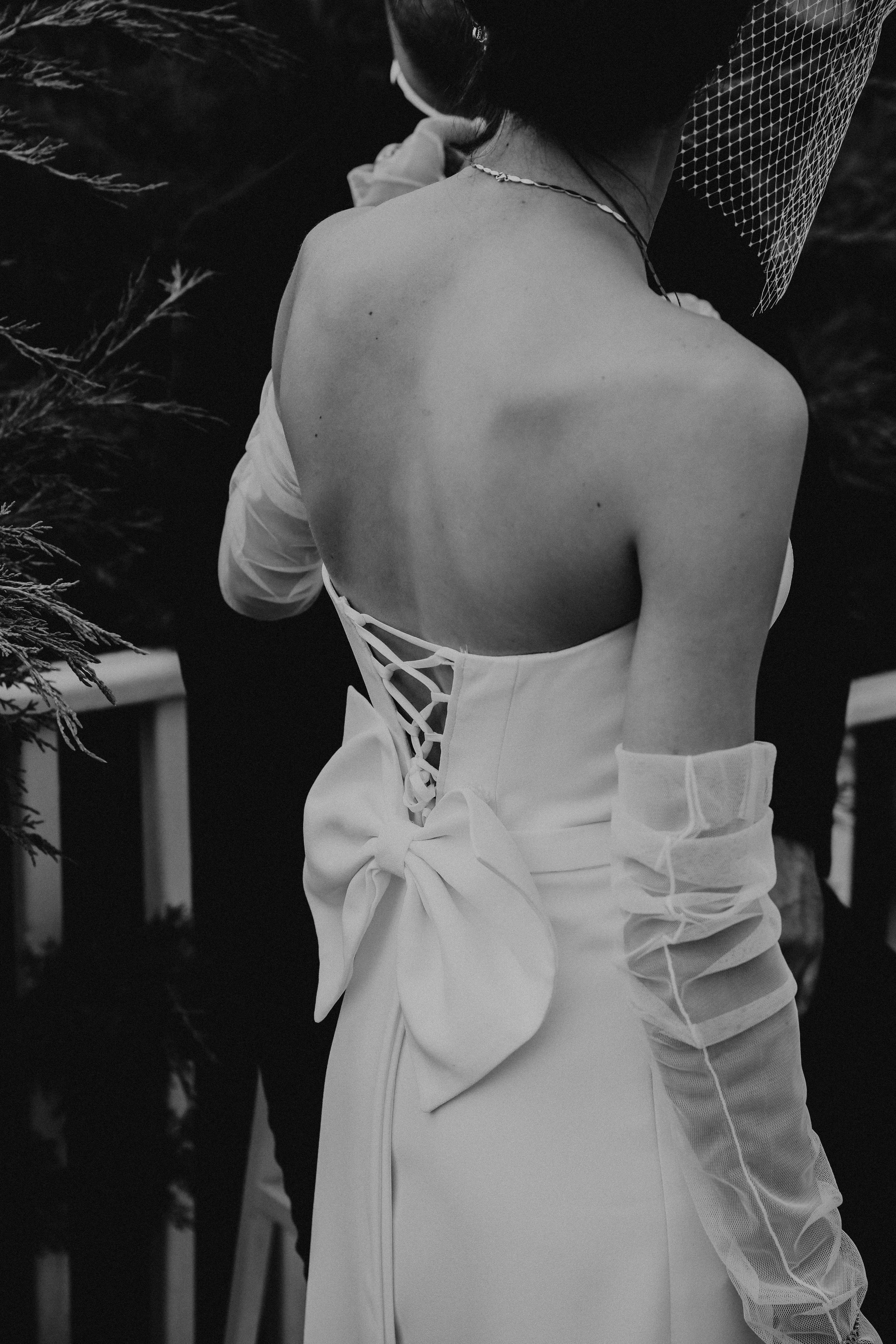 A woman wearing a strapless white dress with a lace-up back and large bow, along with a netted veil and long, sheer glove. She is seen from behind, with her hair styled in a bun, outdoors near some foliage.