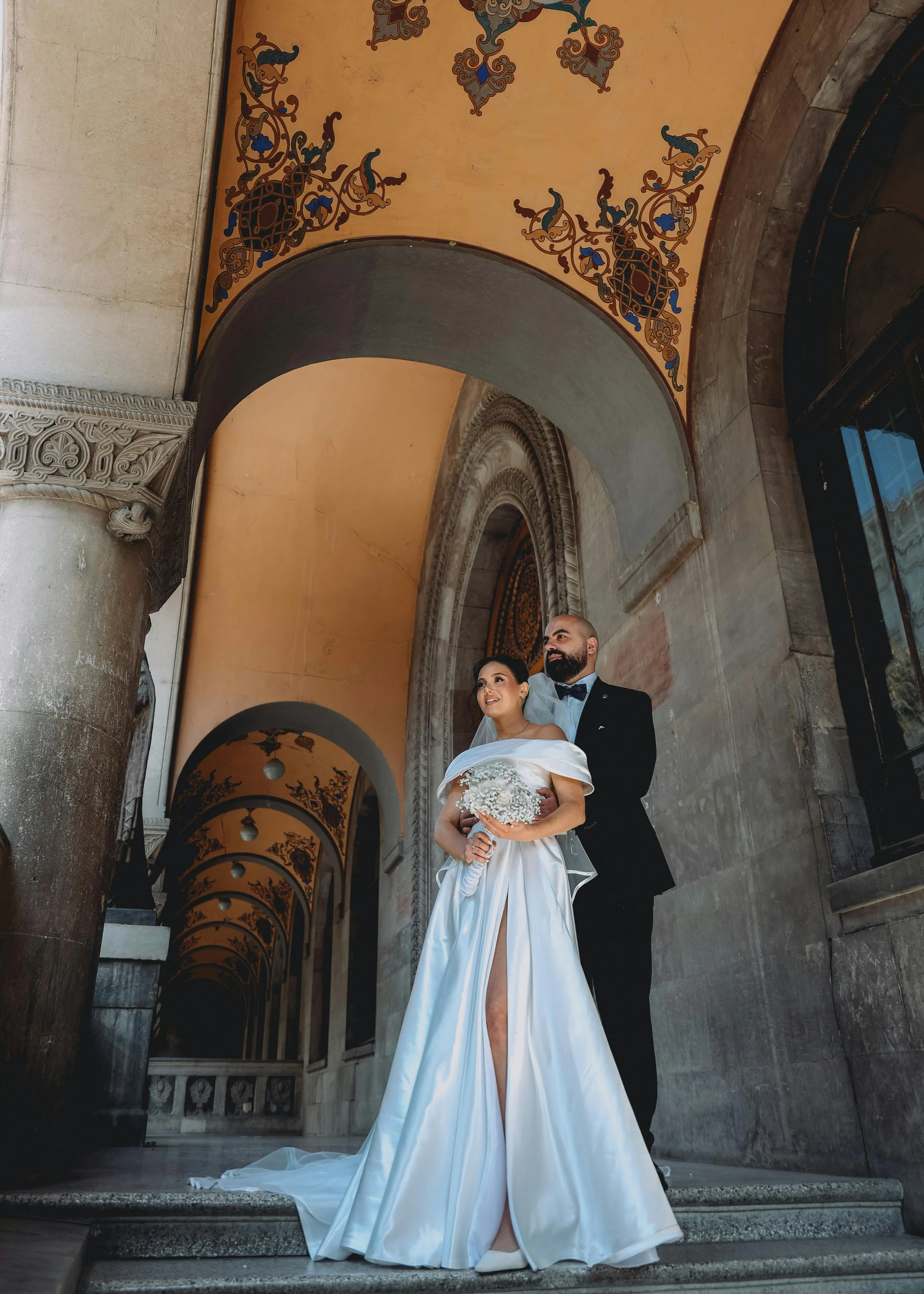 A newlywed couple dressed in wedding attire stands on steps beneath arched, decorated ceilings in a historic building.